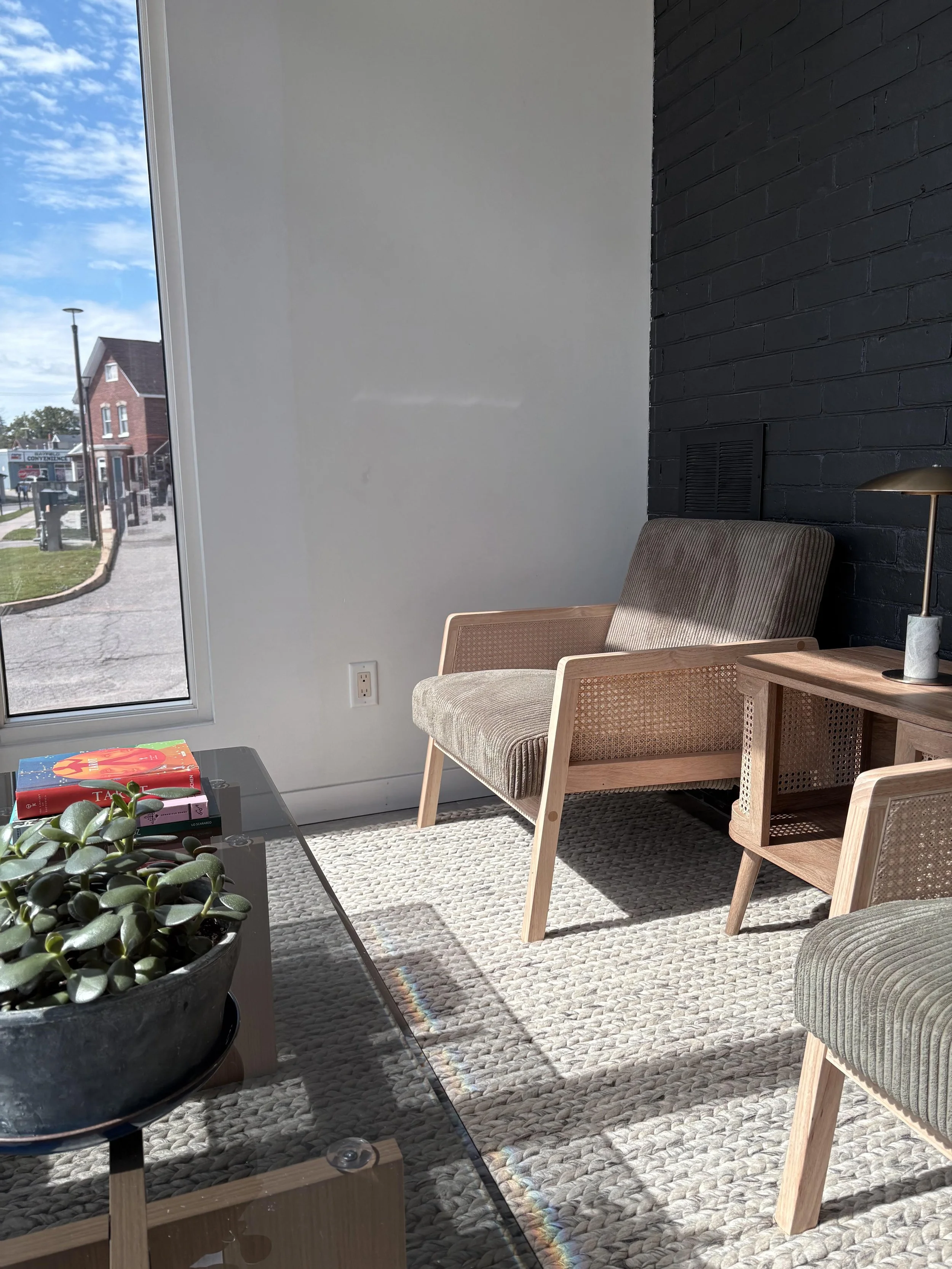 A cozy indoor reading or sitting area near a large window with a view of a street scene, including a brick house, a lamppost, and trees. The area features a beige armchair, a wooden side table, a black coffee table with books and a plant, and a textured rug on the floor. The wall has a black brick accent, and sunlight streams through the window.