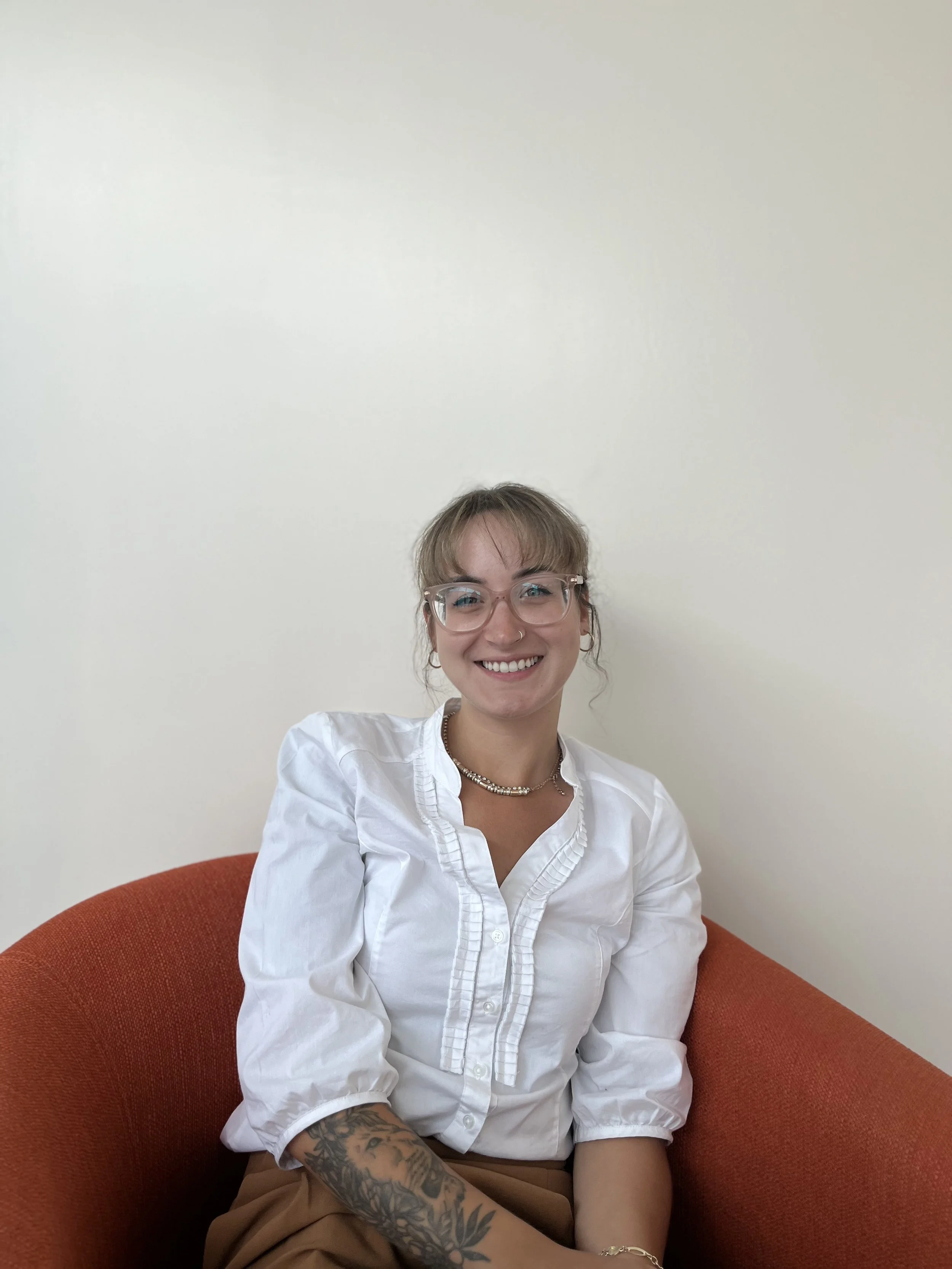 A woman with glasses and tattoos smiling, sitting on a red couch against a plain white wall.