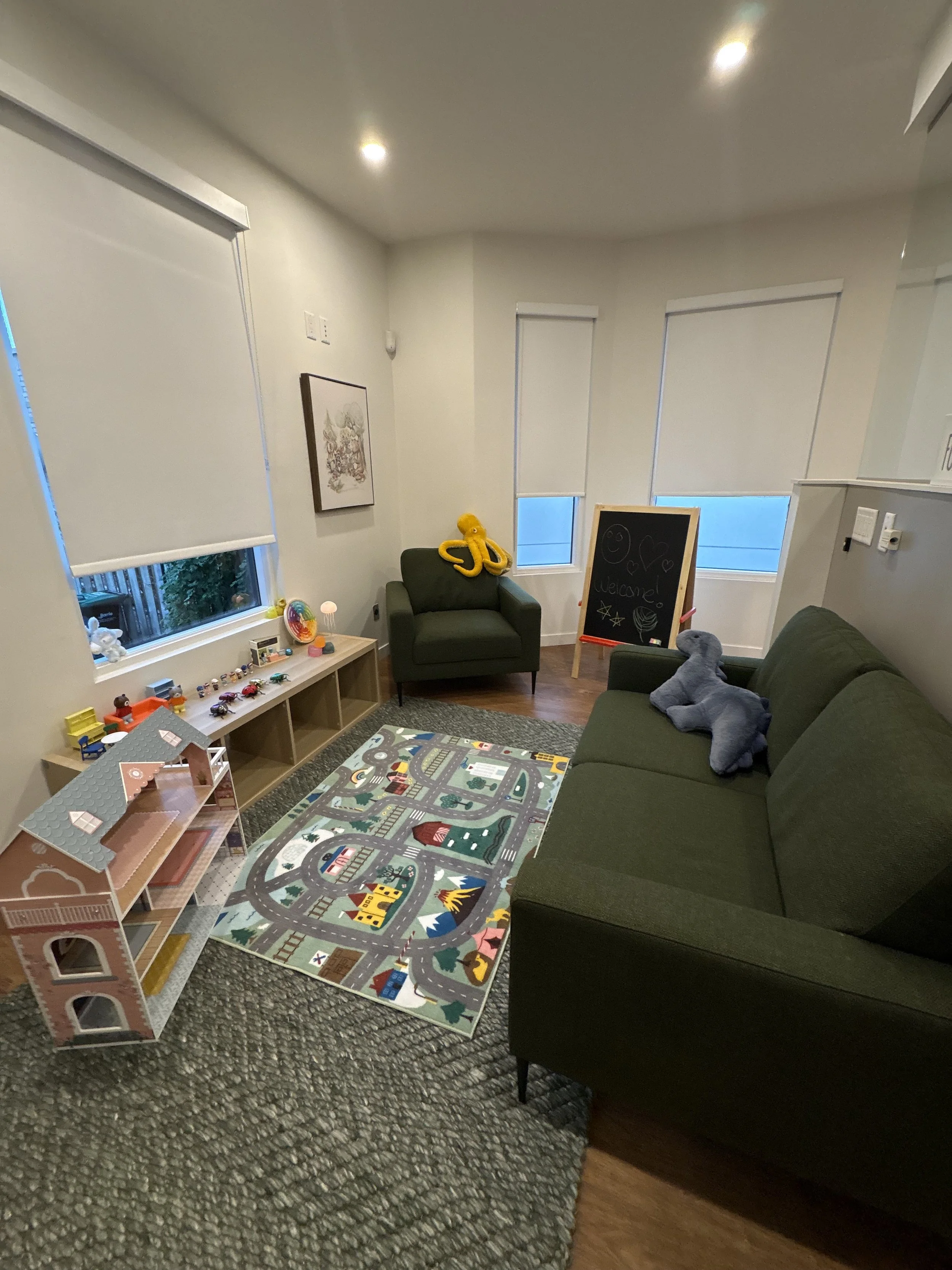 Living room with green sofa, children’s toys, a playhouse, a colorful play mat, a blackboard with drawings, and windows with blinds.
