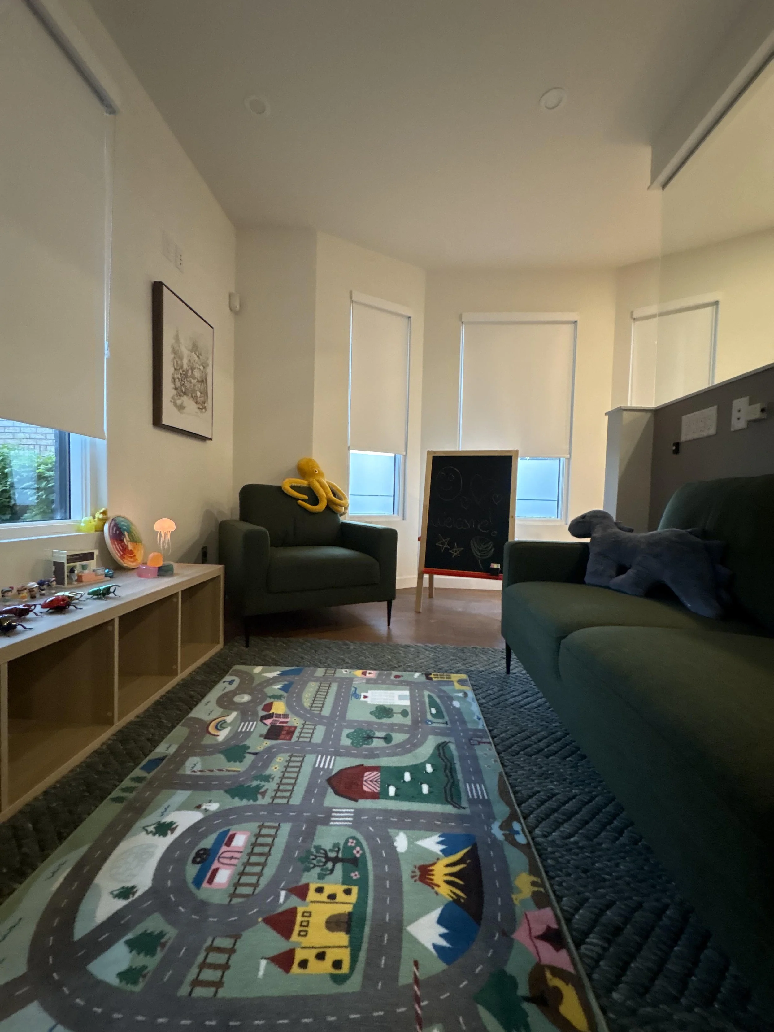 Living room with large colorful road-themed playmat on wooden floor, dark green armchair, blackboard, window with blinds, and small decorative items on a table.