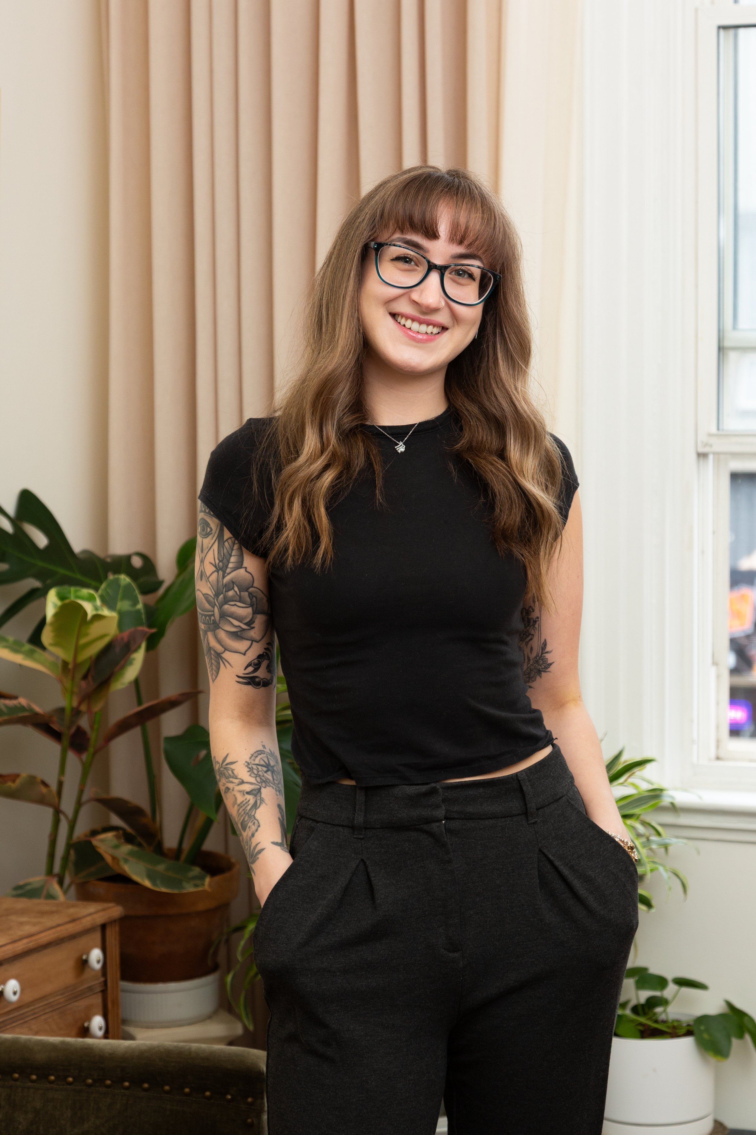 A young woman with glasses, long brown wavy hair, and tattoos on her arm, wearing a black t-shirt and dark pants, standing indoors near houseplants, smiling at the camera.