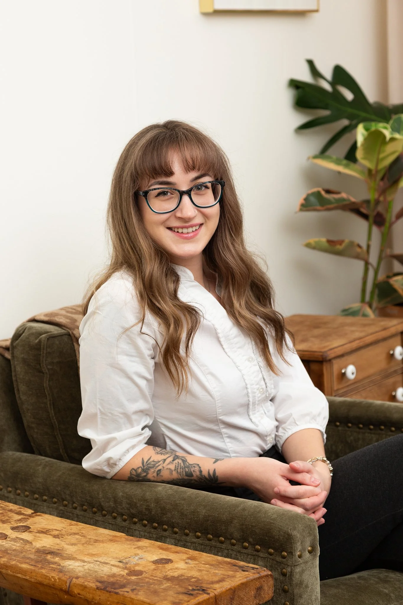 A young woman with long, wavy, light brown hair, wearing glasses and a white button-up shirt, sitting on a brown upholstered chair. She is smiling and has a tattoo on her right forearm. The background includes a plant with green and yellow leaves and a light-colored wall.