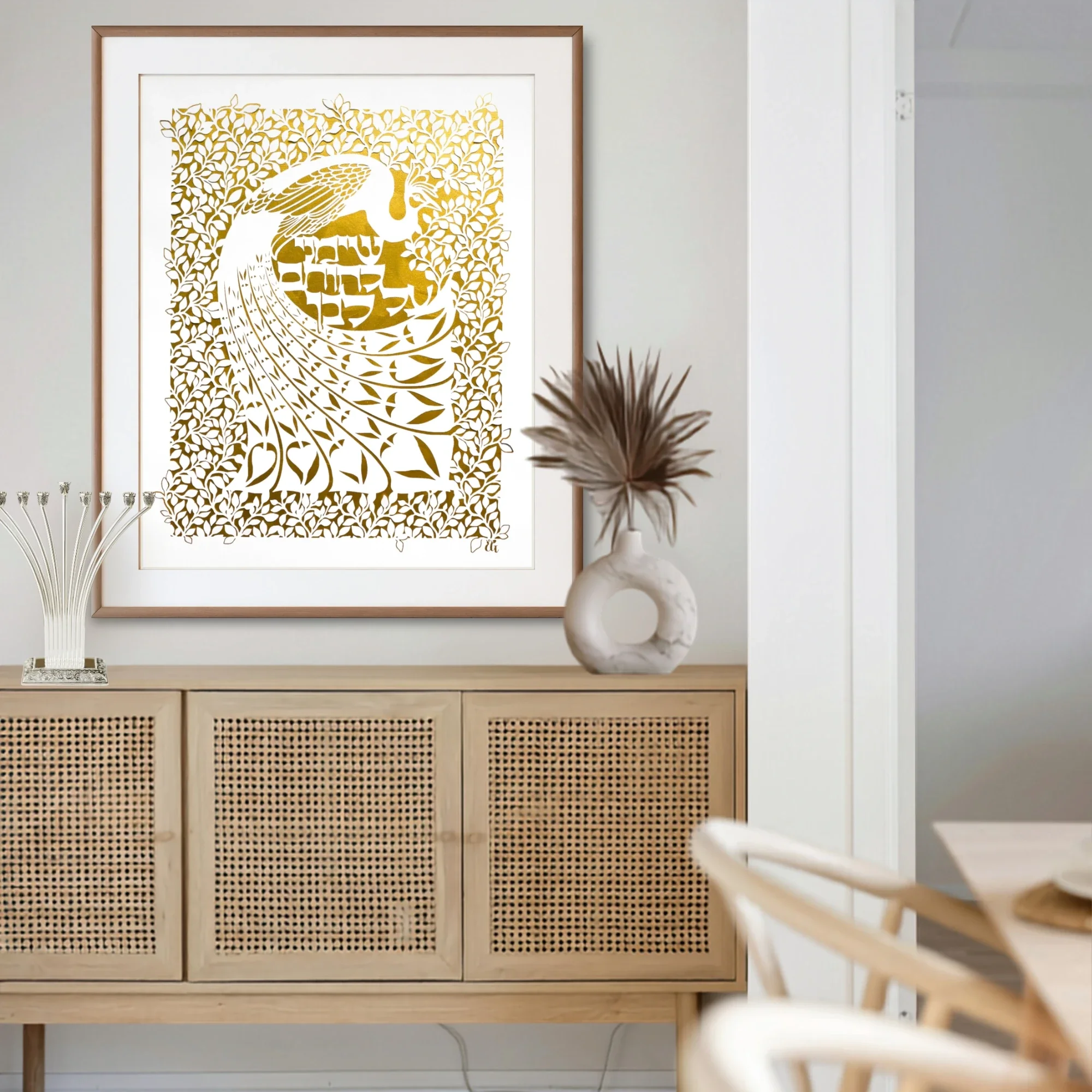 A framed papercut artwork of a peacock featuring intricate botanical designs and Hebrew, displayed with a wooden furniture in a bright dining room.