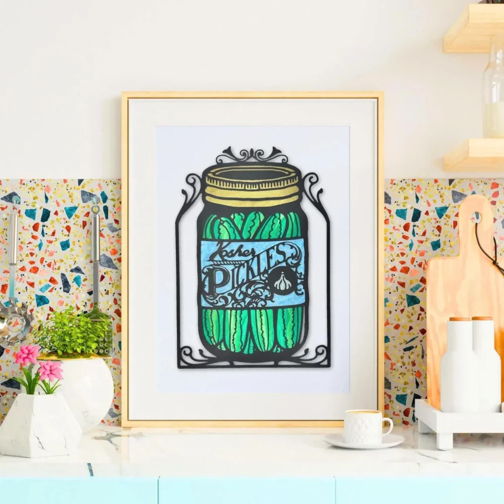 Framed pickle jar papercut art framed and leaning against a bright kitchen backsplash with plants and countertop accessories.