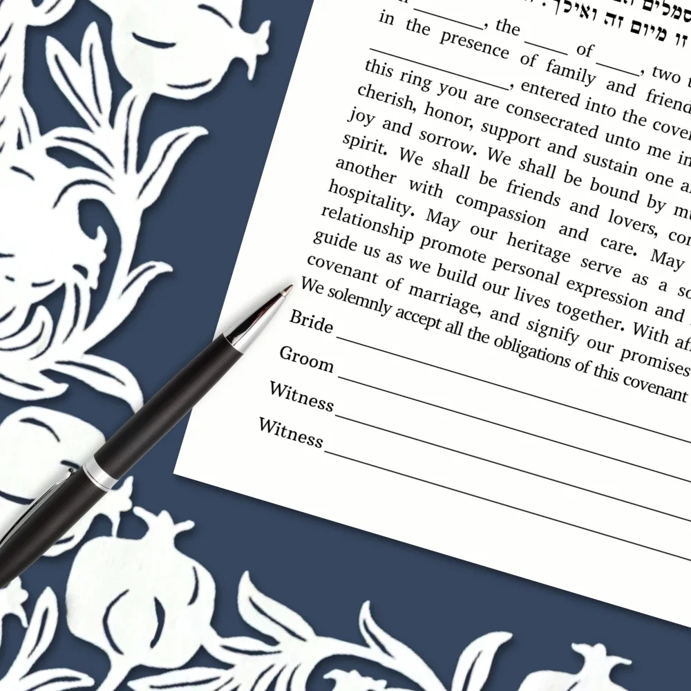 close up of white and blue papercut ketubah with pomegranates, and a pen laying across the text, ready to sign