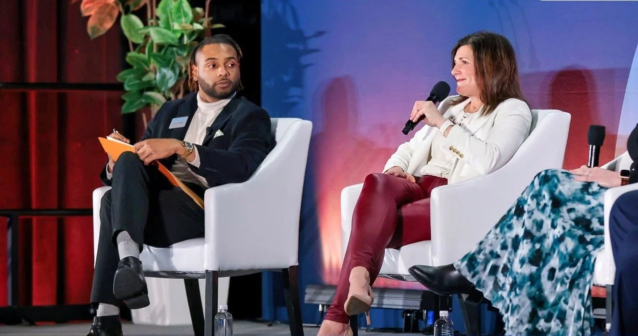Three people sitting in white chairs on a stage, participating in a panel discussion. The woman on the right is holding a microphone and speaking, while the man on the left is holding a notepad and listening. The background features a red and blue backdrop, with some plants visible.