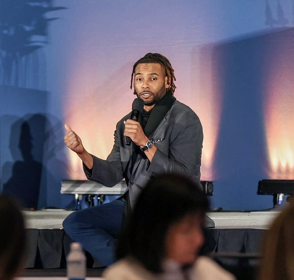 Keenan Medlock speaking to a live audience with a microphone onstage.