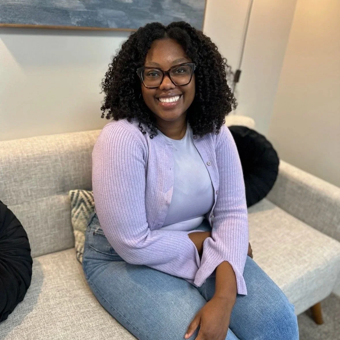 Smiling woman with curly hair and glasses sitting on a light-colored sofa