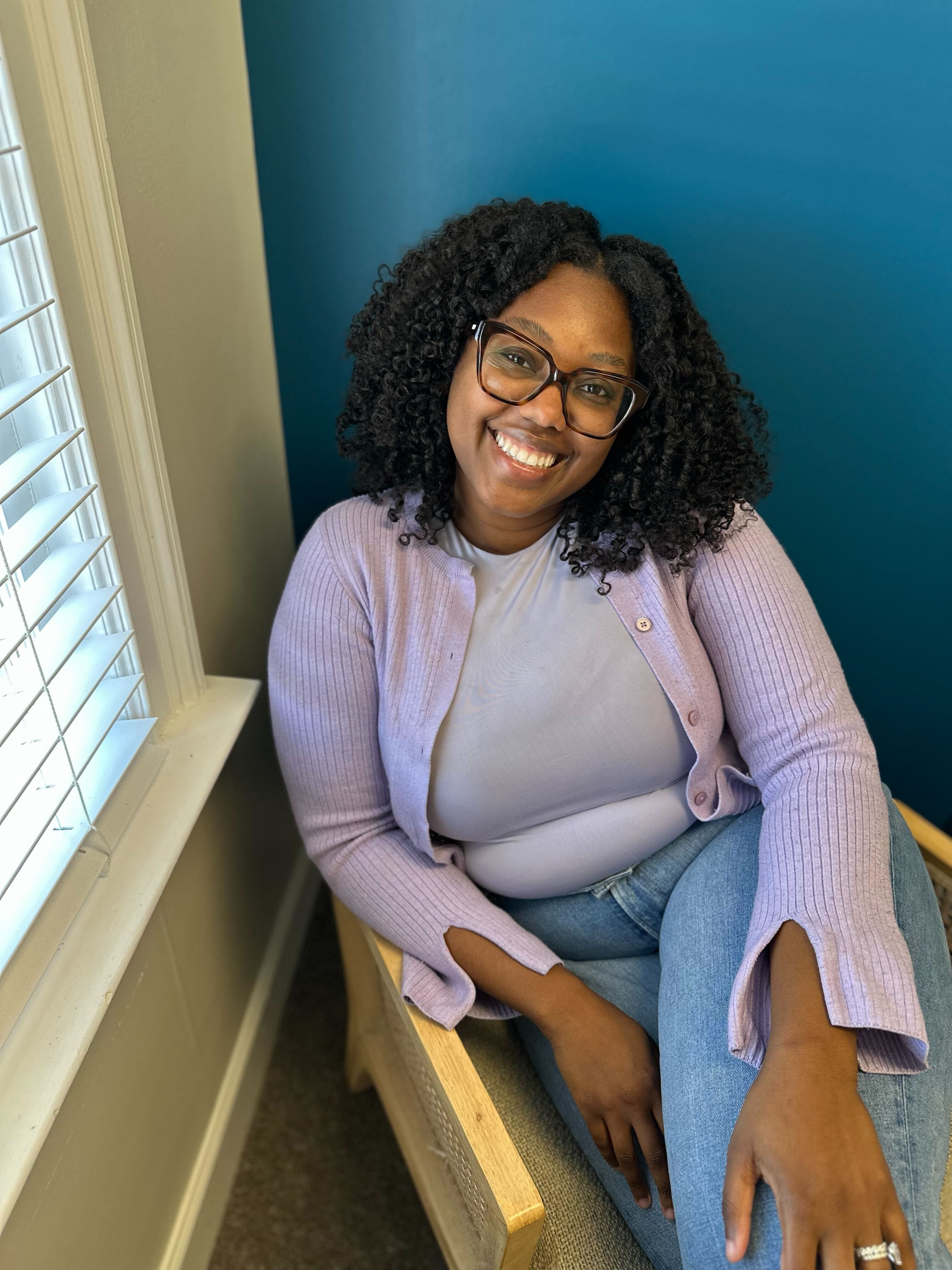 Sydney Christian, LPC is sitting in a tan chair with a dark blue wall behind her. She has curly shoulder length hair and is wearing glasses, a purple top and cardigan, and jeans. She is smiling