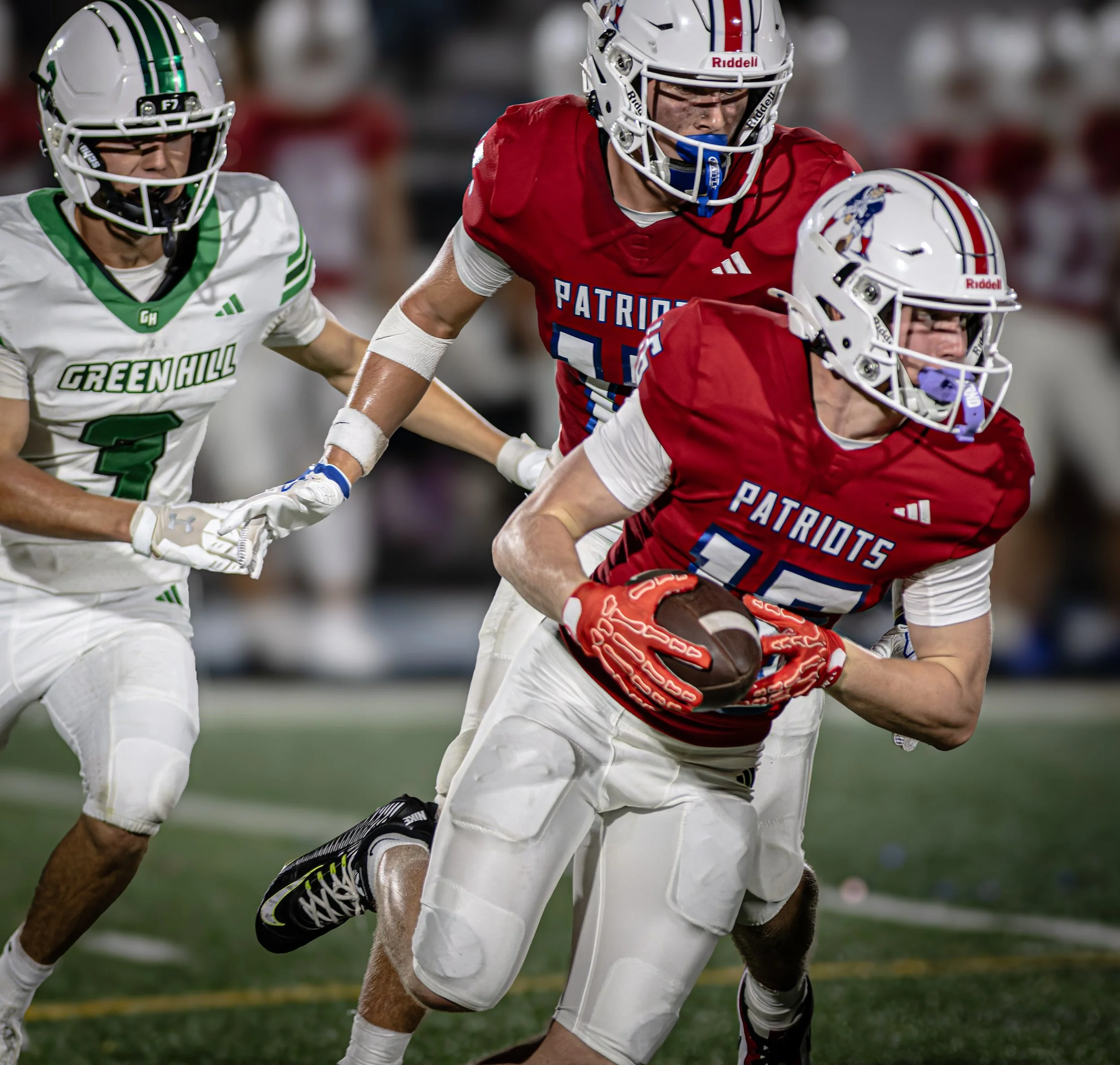 Two football players, one in a red jersey with 'Patriots' written on it and the other in a white jersey with green accents and 'Green Hill' written on it, are engaged in a game. The player in red is holding a football and is being tackled by the play