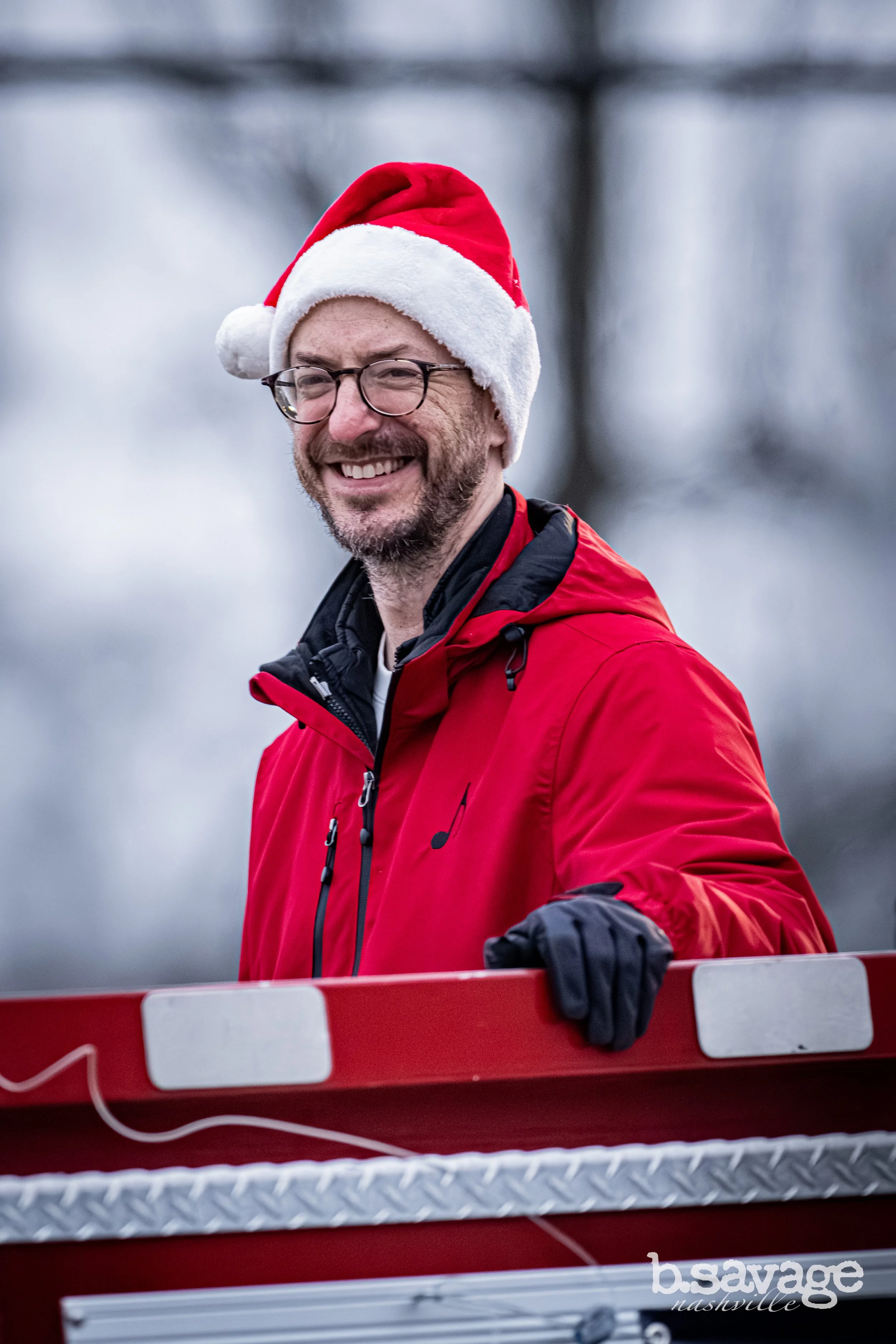 A smiling man wearing a Santa hat and glasses stands behind a red and white barricade, outdoors on a cloudy day.