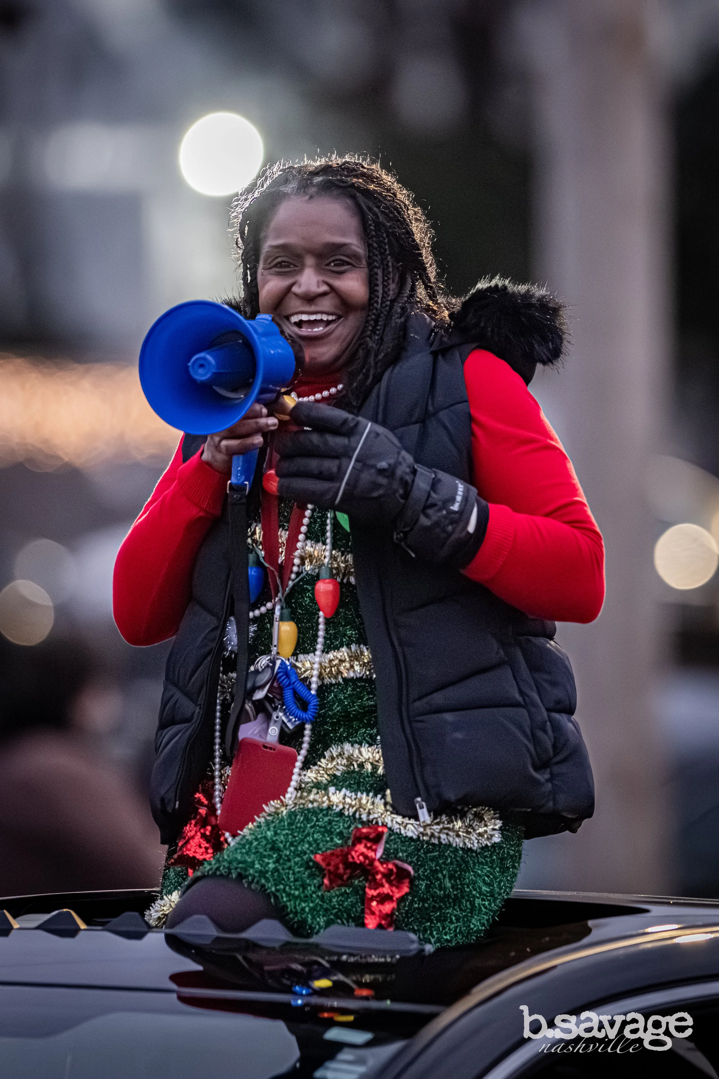 A woman smiling and speaking into a blue megaphone, dressed in a festive green and red Christmas outfit with decorations, standing outdoors during a celebration or parade.