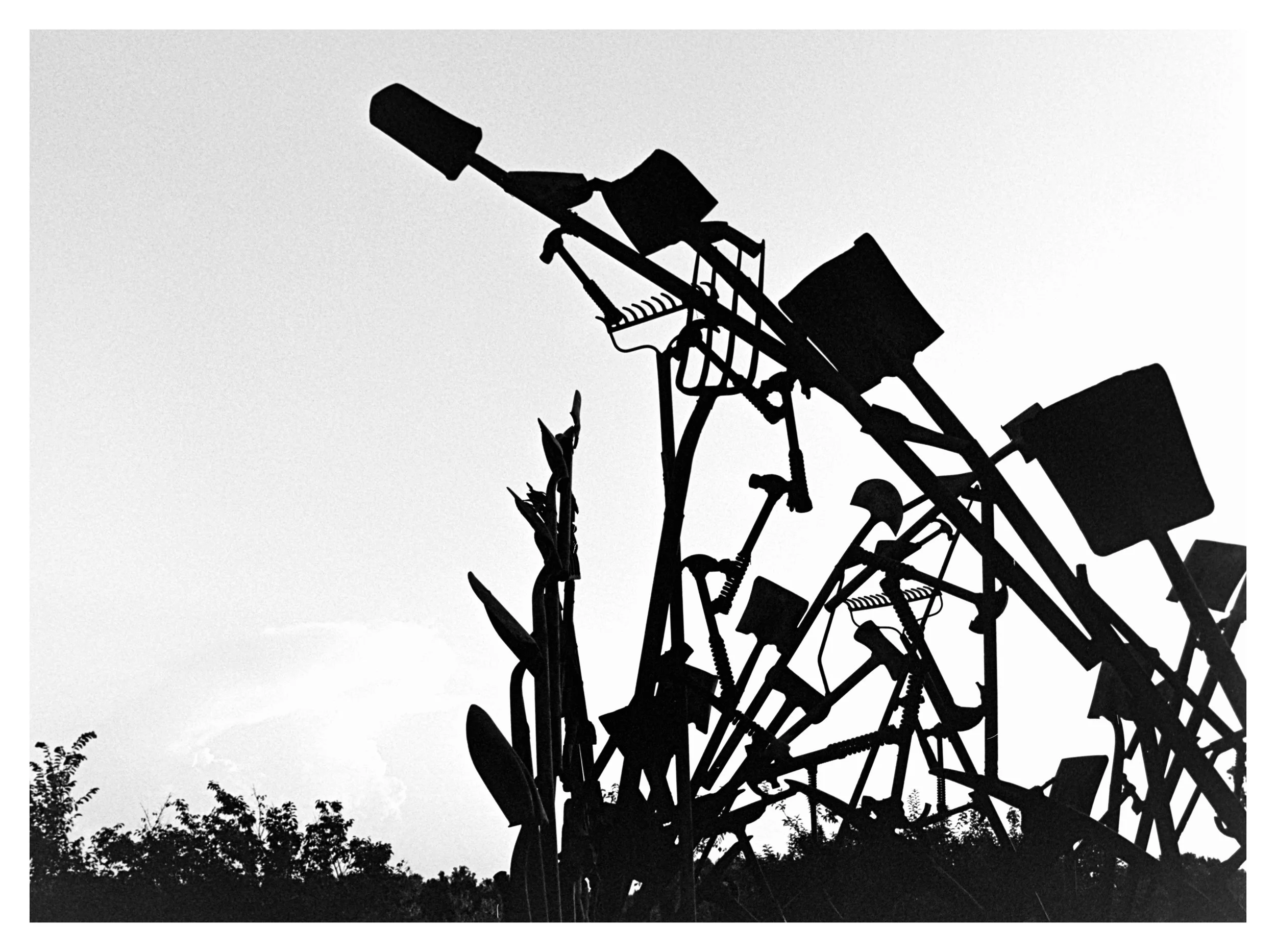 Silhouette of garden tools, including a rake, shovel, and hoes, arranged against a light sky with trees in the foreground.
