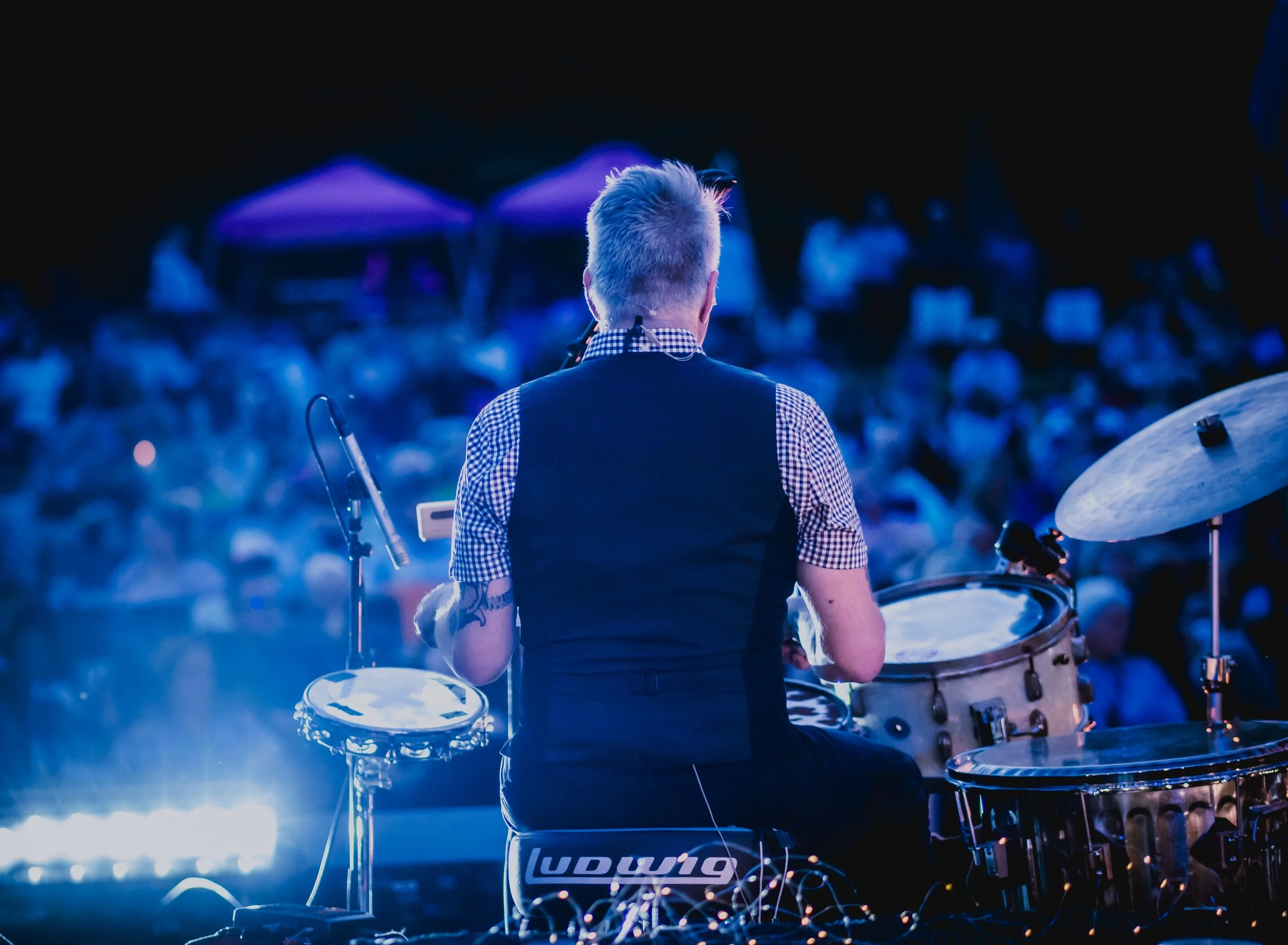 Back of a drummer with gray hair playing drums on stage at night, illuminated by blue lights with umbrellas in the background.