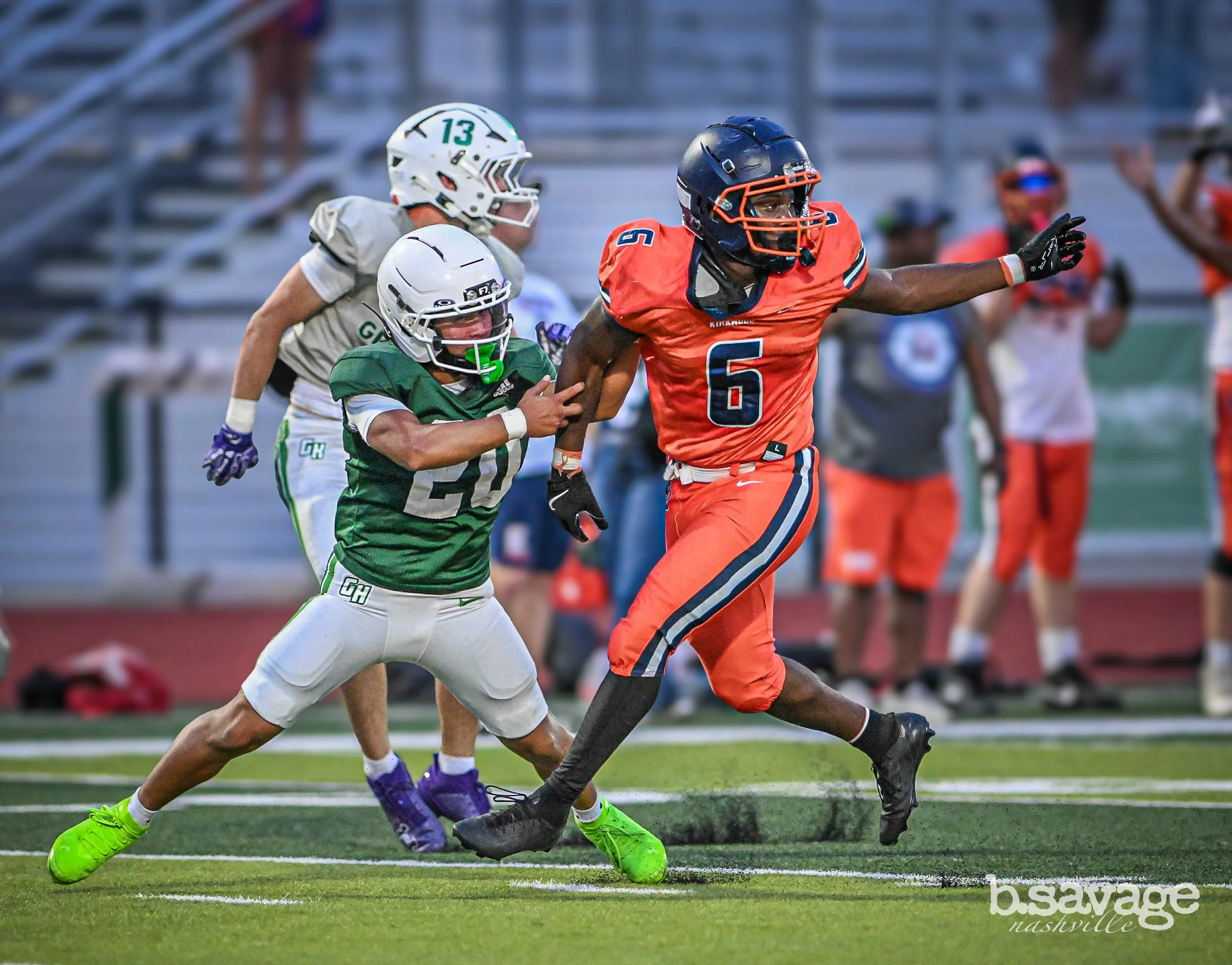 Two football players in action during a game, one in a green uniform and the other in an orange uniform, with a football helmet and pads, on a football field with spectators in the background.