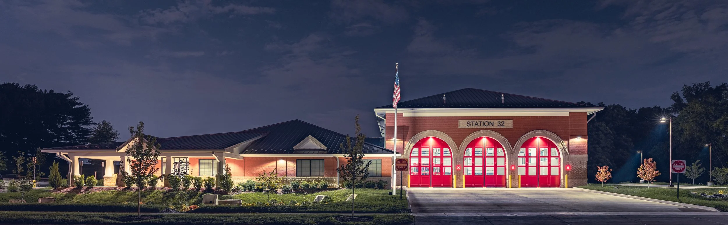 A fire station with red doors, a sign reading 'Station 32', an American flag, and surrounding trees illuminated at night.