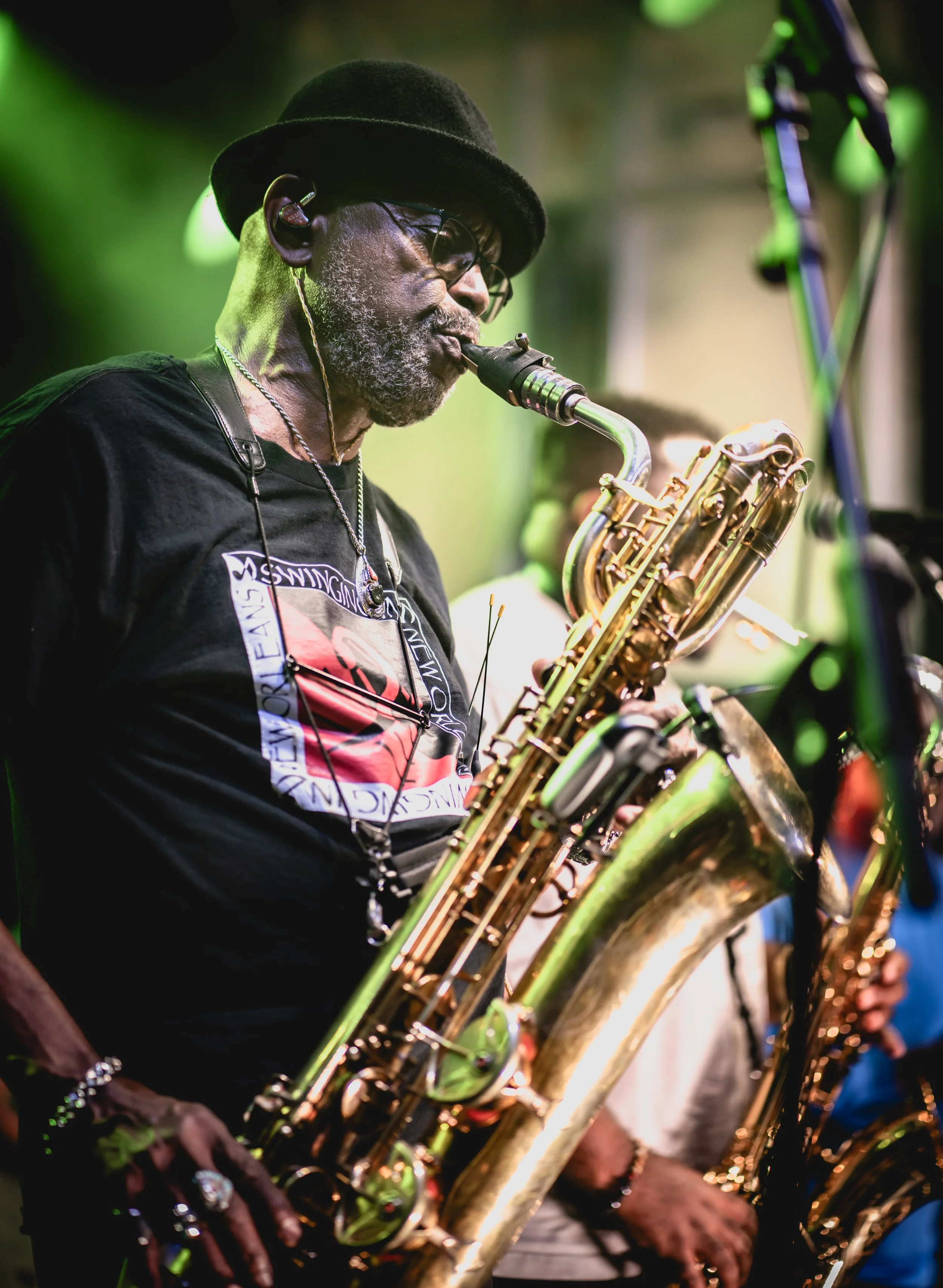 A musician plays the saxophone at a live performance, wearing a black hat, glasses, and a black T-shirt with a graphic design, with other band members partially visible in the background.