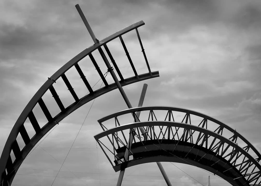 Black and white photo of a fallen amusement park ride, possibly a roller coaster, with bent rails and supports against a cloudy sky.