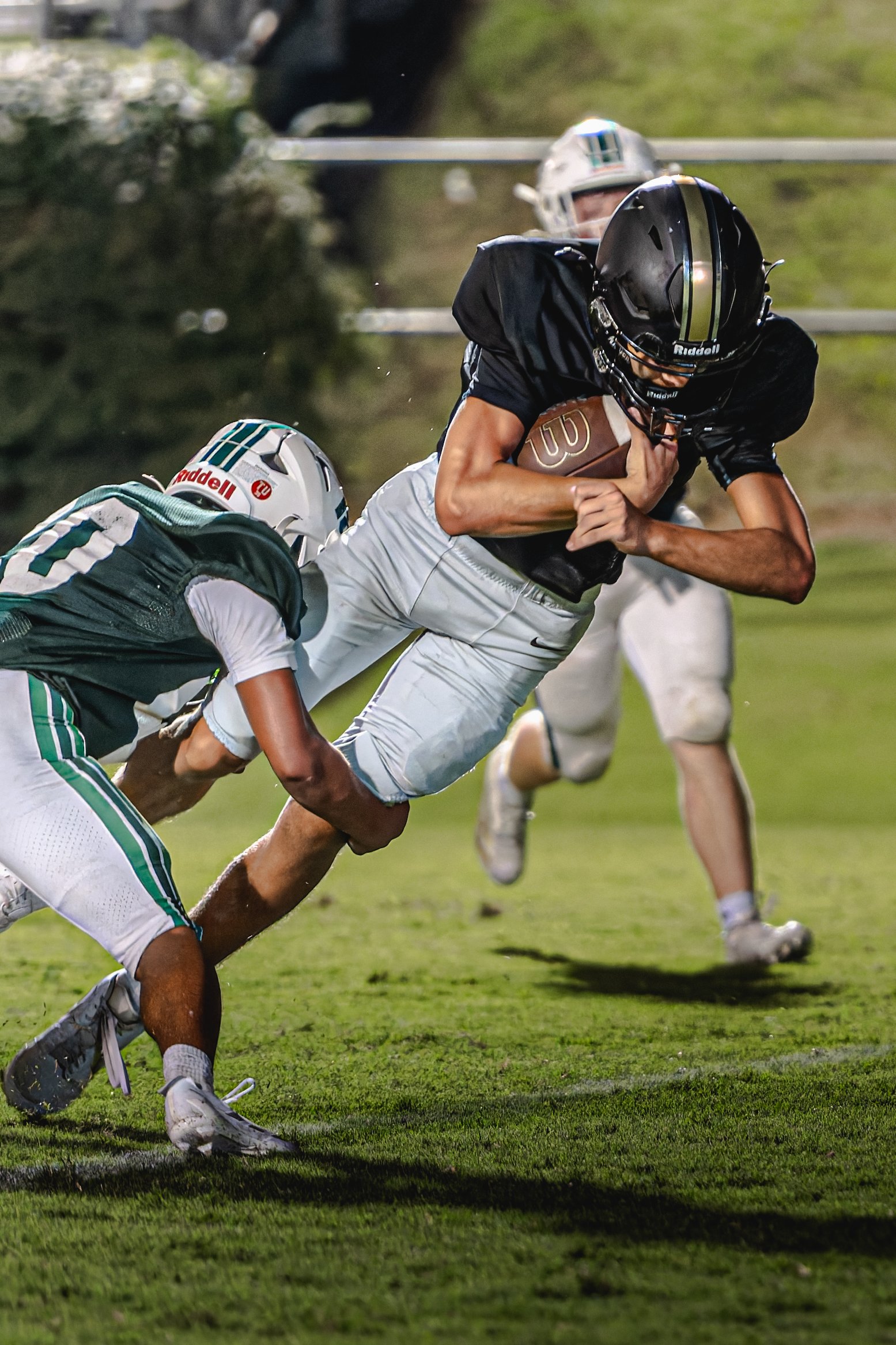 A football player carrying the ball runs past opponents during a night game on a grassy field.
