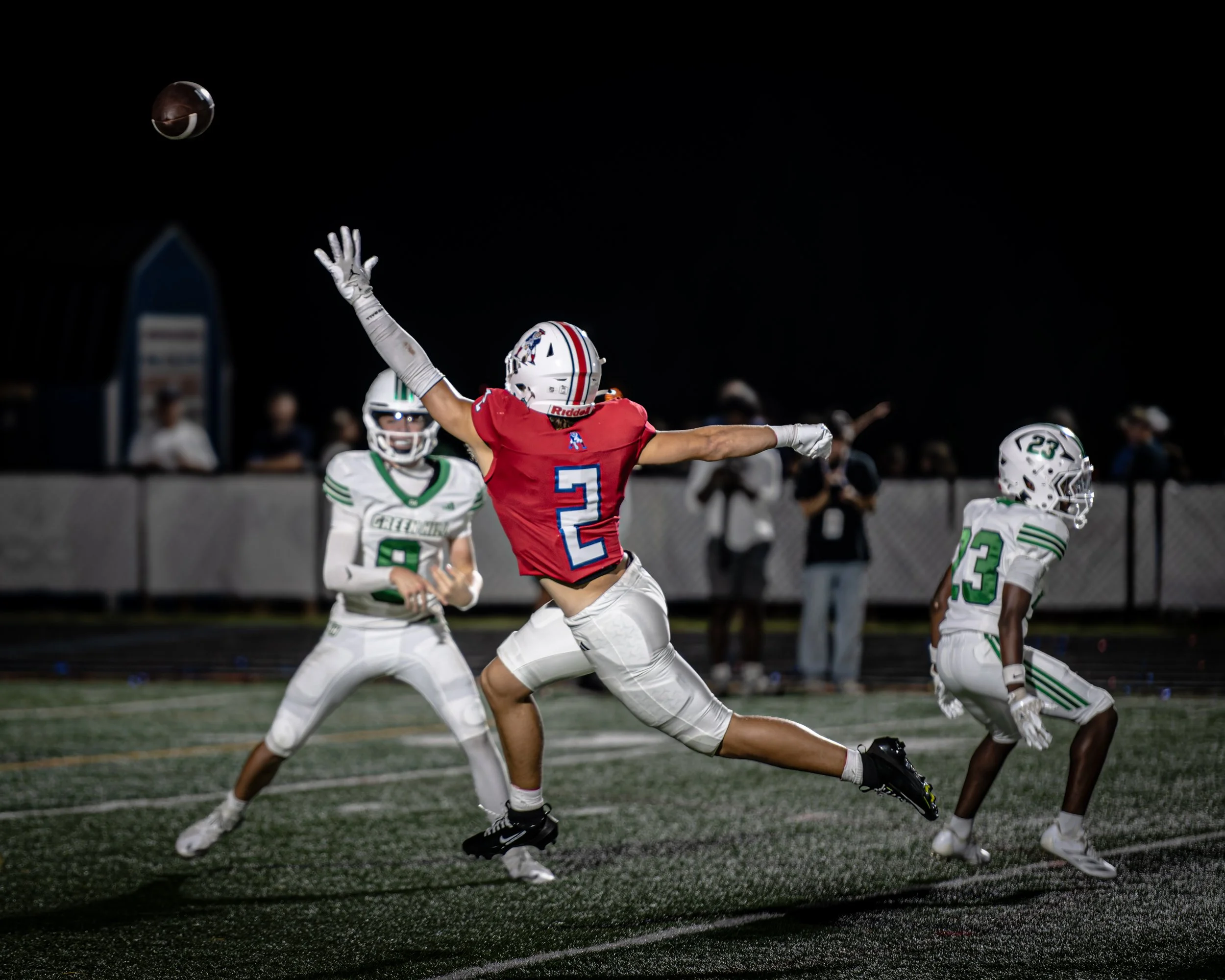 Football players on the field at night, with one player in a red jersey jumping to catch the ball while others in white jerseys look on.