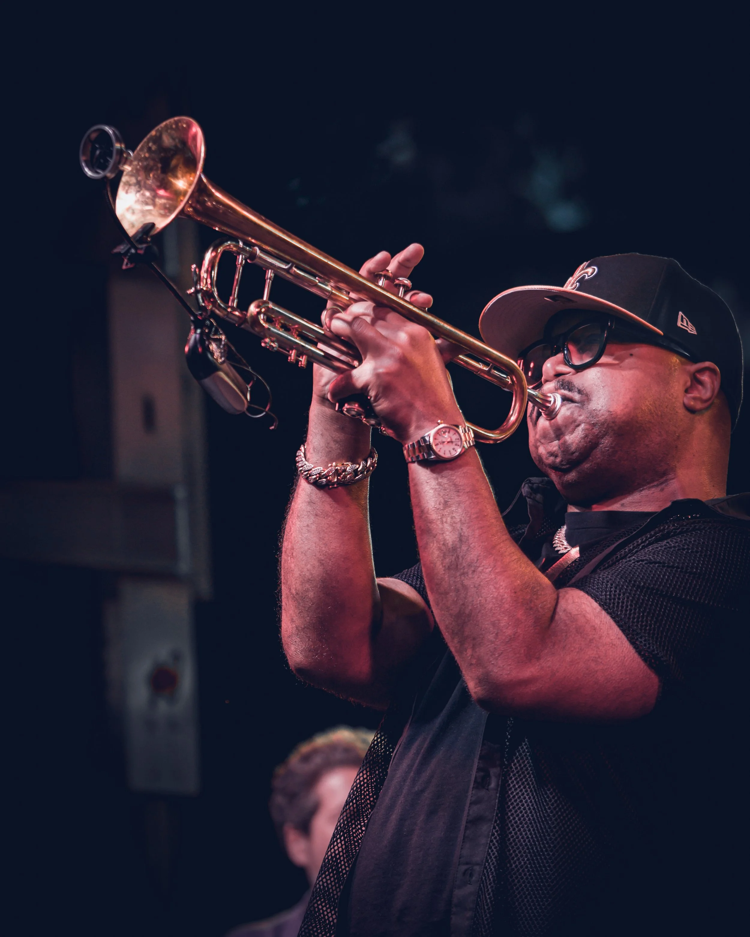 A man wearing a black cap, glasses, and a wristwatch is playing a gold trumpet. The background is dark, and there is another person slightly visible in the background.