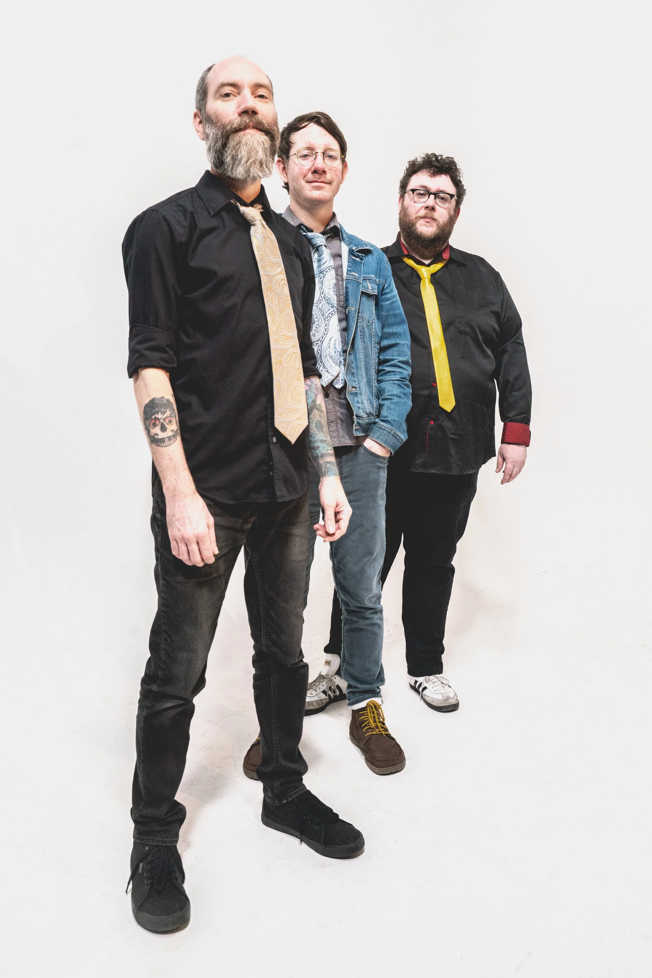Three men standing against a plain white background, each dressed in casual attire with one wearing a tie, one wearing a denim jacket, and the third in a black shirt with a yellow tie. The man on the left has a beard and tattoos on his forearm, the m