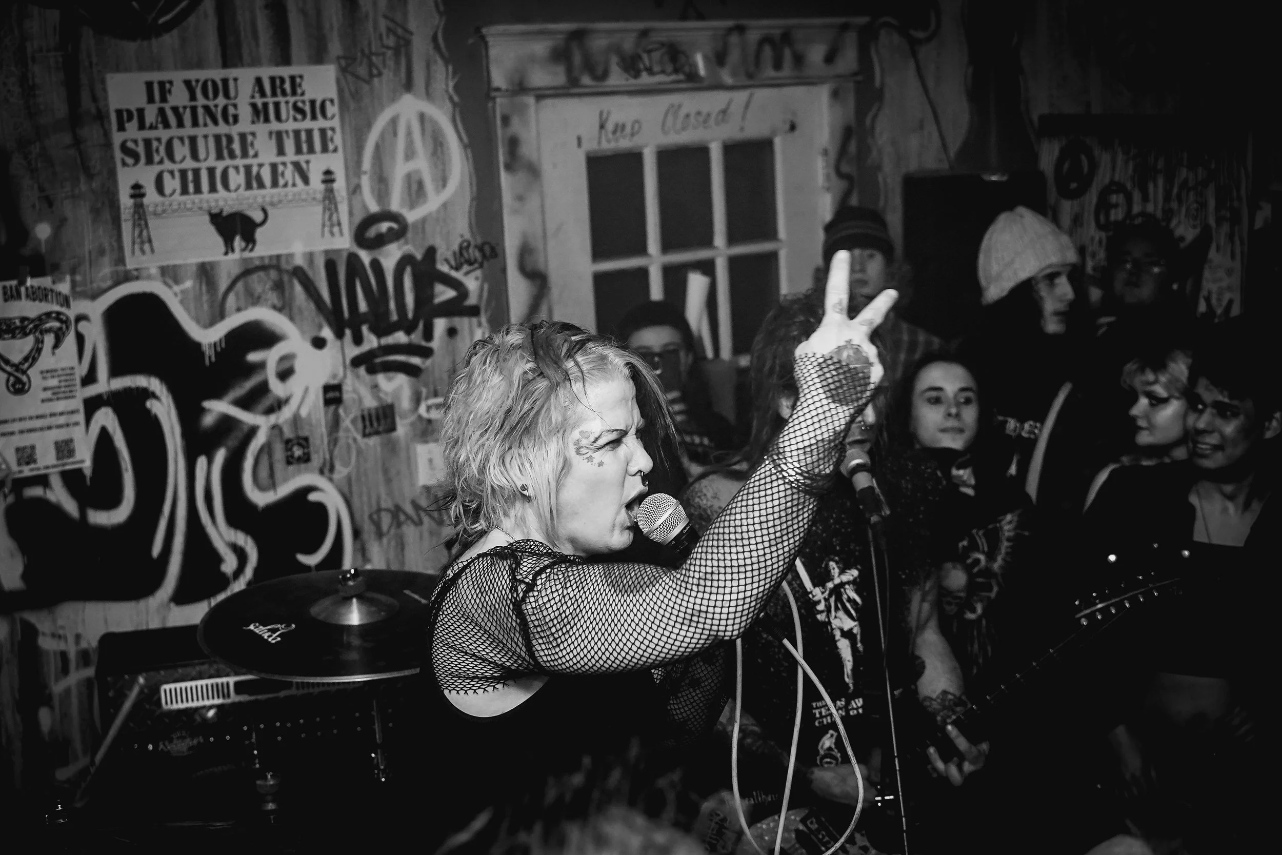 A woman with blonde hair singing into a microphone and playing guitar in a dimly lit room decorated with graffiti and posters, with a group of people in the background.