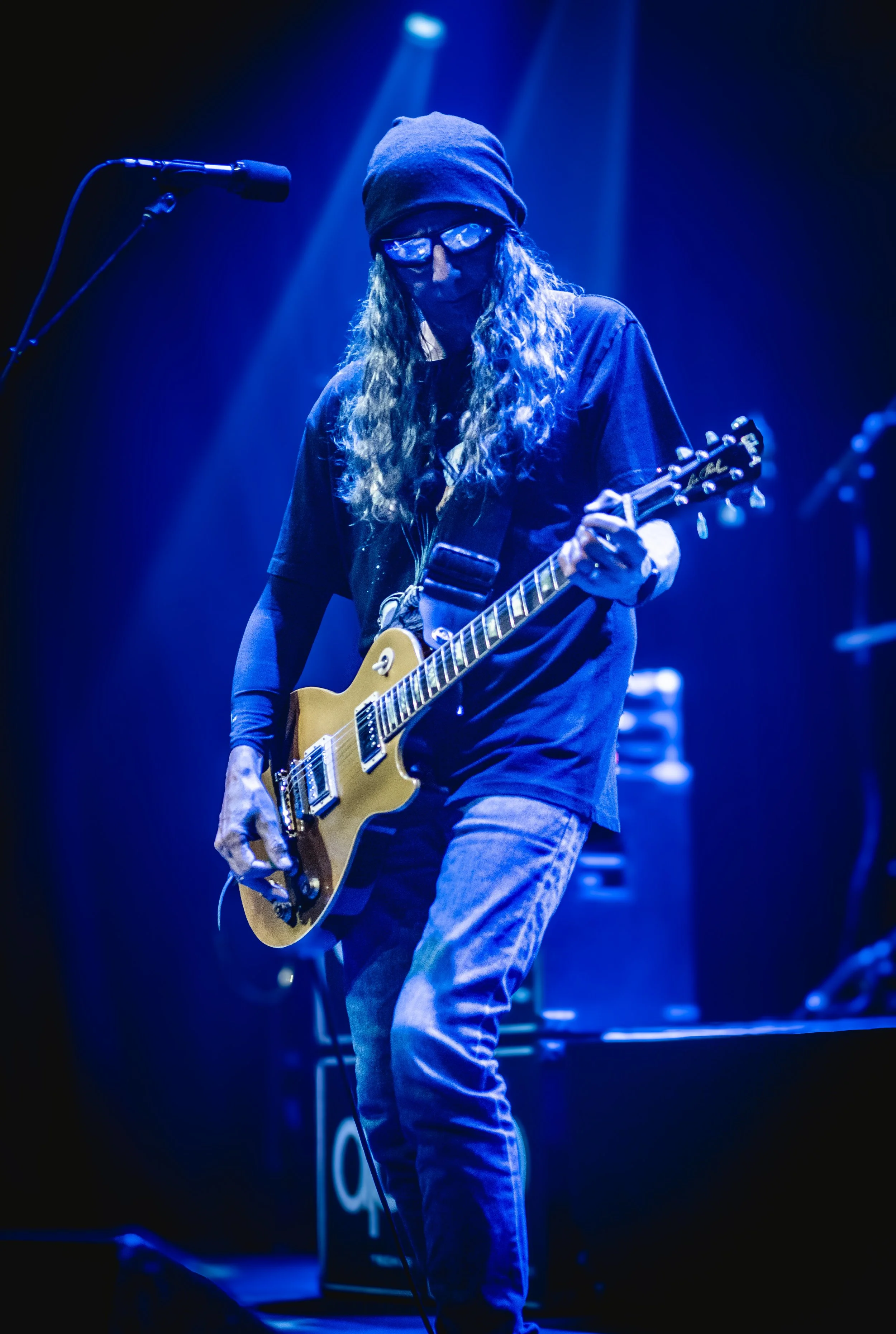 A musician with long curly hair, wearing a beanie, sunglasses, and dark clothing, playing an electric guitar on stage illuminated by blue lights.