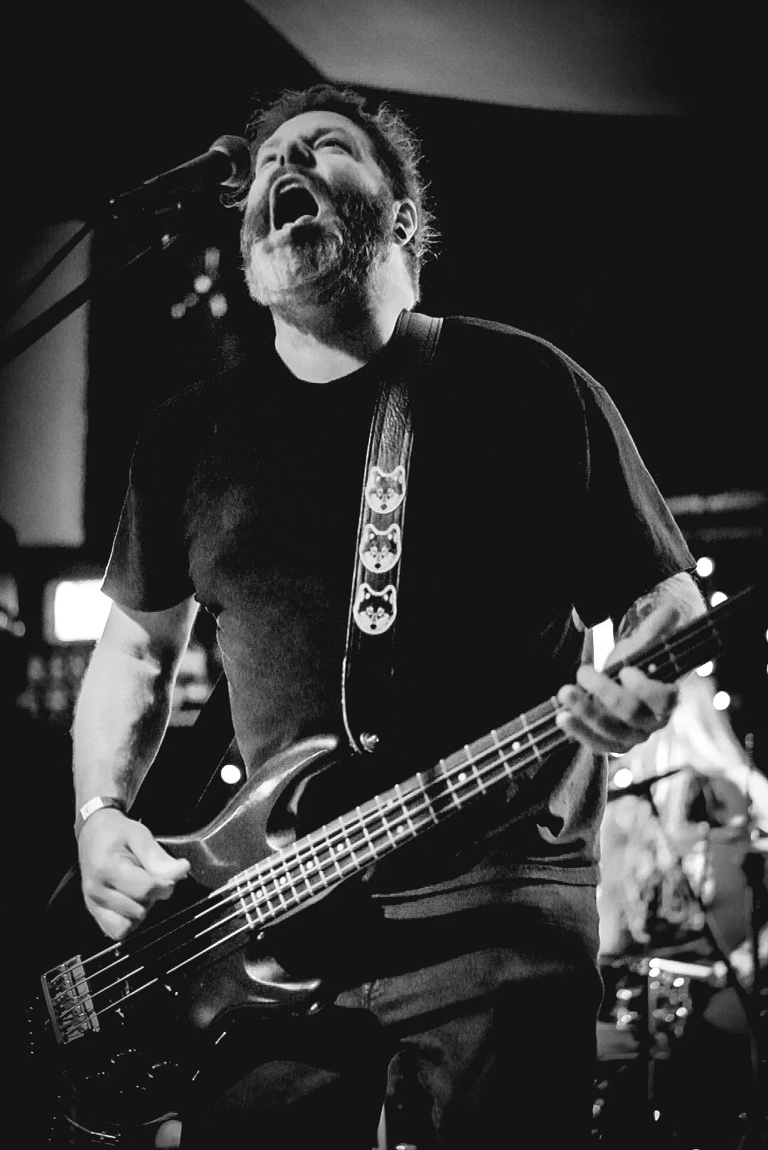 A black and white photo of a bearded man playing an electric bass guitar with his mouth open, singing or shouting passionately during a performance. He is wearing a black t-shirt with a guitar strap decorated with cartoon-like face designs.