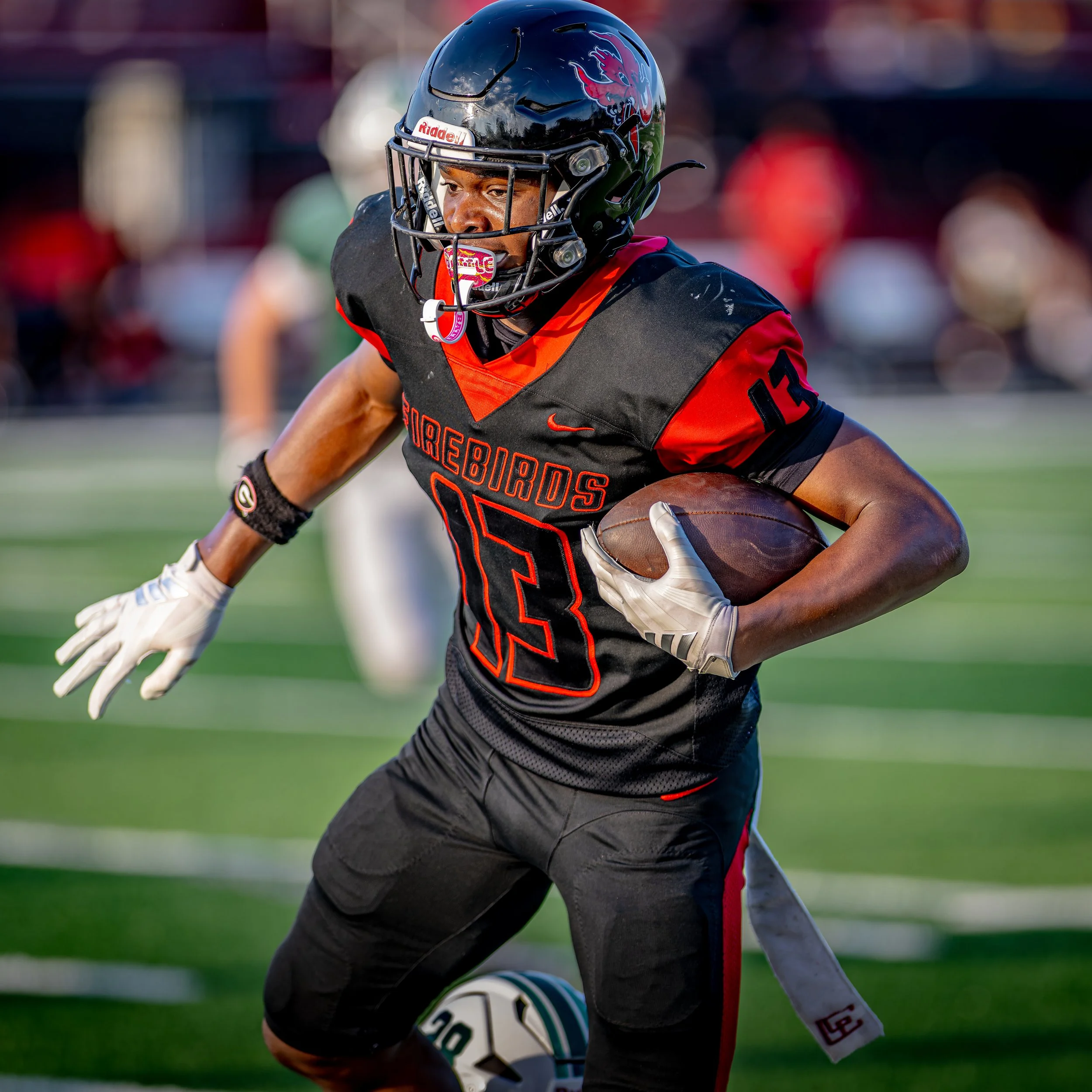 A football player in black and red uniform crouching on the field, holding a football, wearing a black helmet with a red dragon logo, and white gloves, during a game with a blurred crowd in the background.