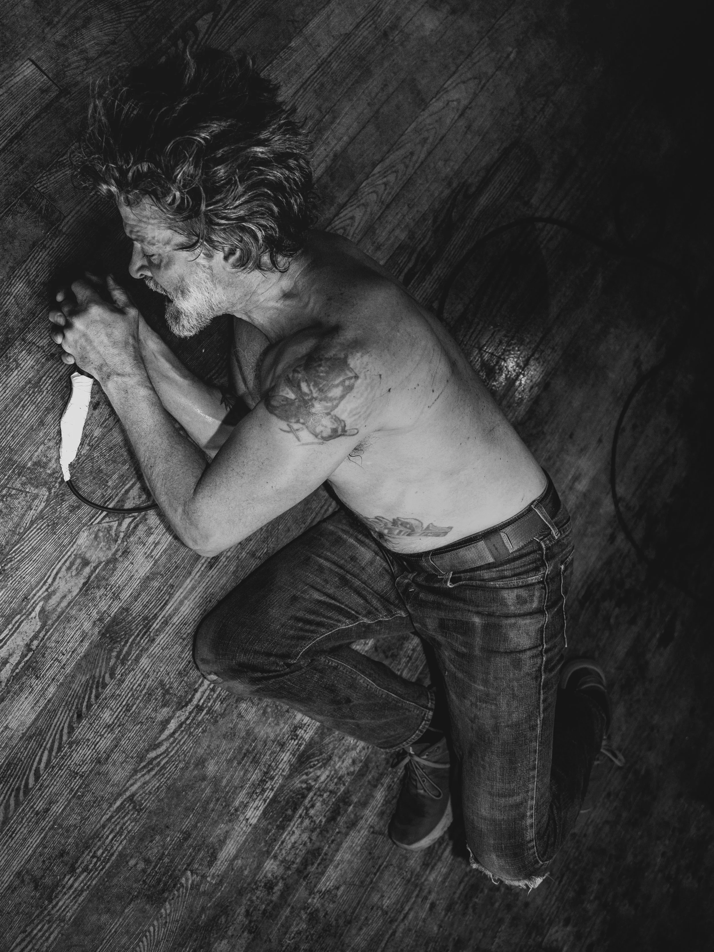 A black and white photo of an older man with long hair and a beard, lying on a wooden floor with his eyes closed, holding a light-colored object in his hands, shirtless with visible tattoos on his arm and side.