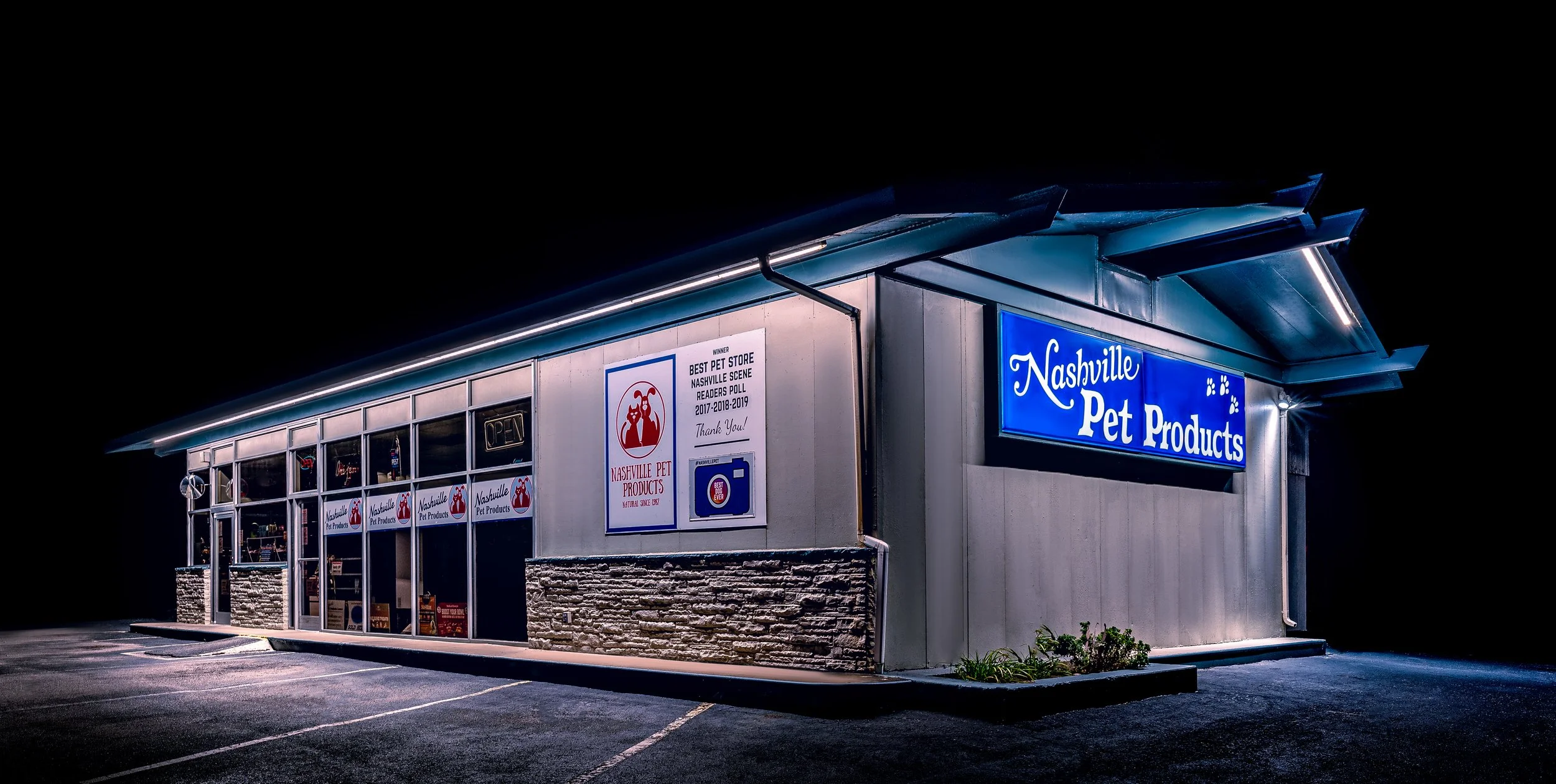 Nighttime exterior view of a Nashville Pet Products store with illuminated signs and windows, showing parking lot with few empty spaces.