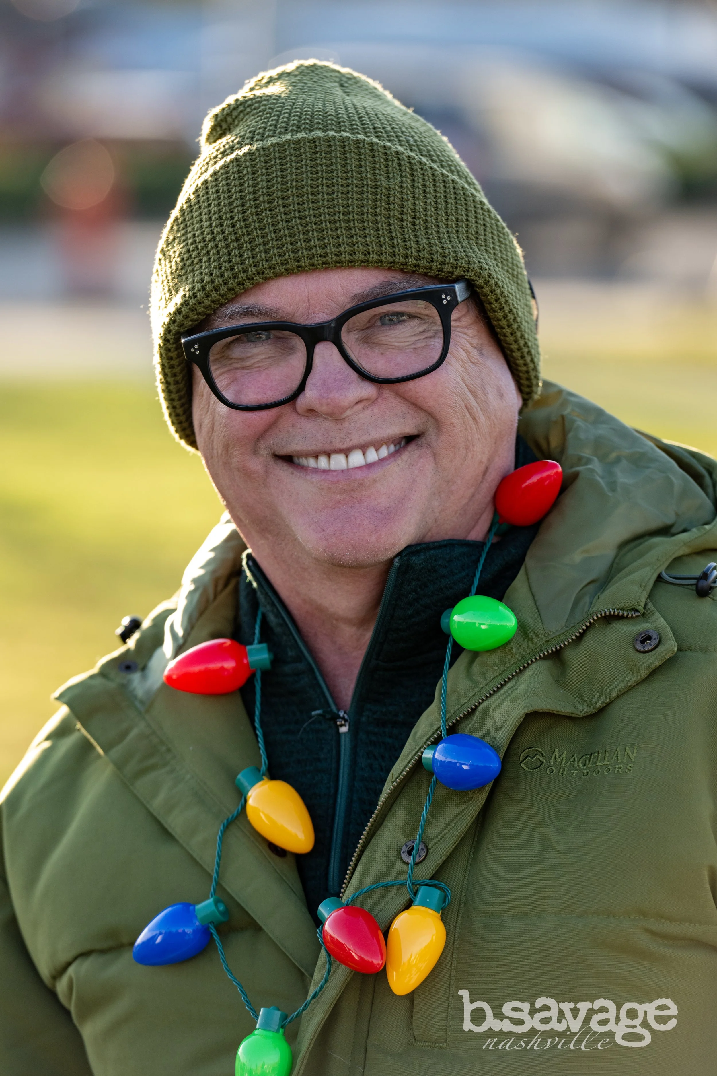A smiling man wearing a green knit beanie, black glasses, a green jacket, and colorful holiday lights around his neck, outdoors with a blurred background.