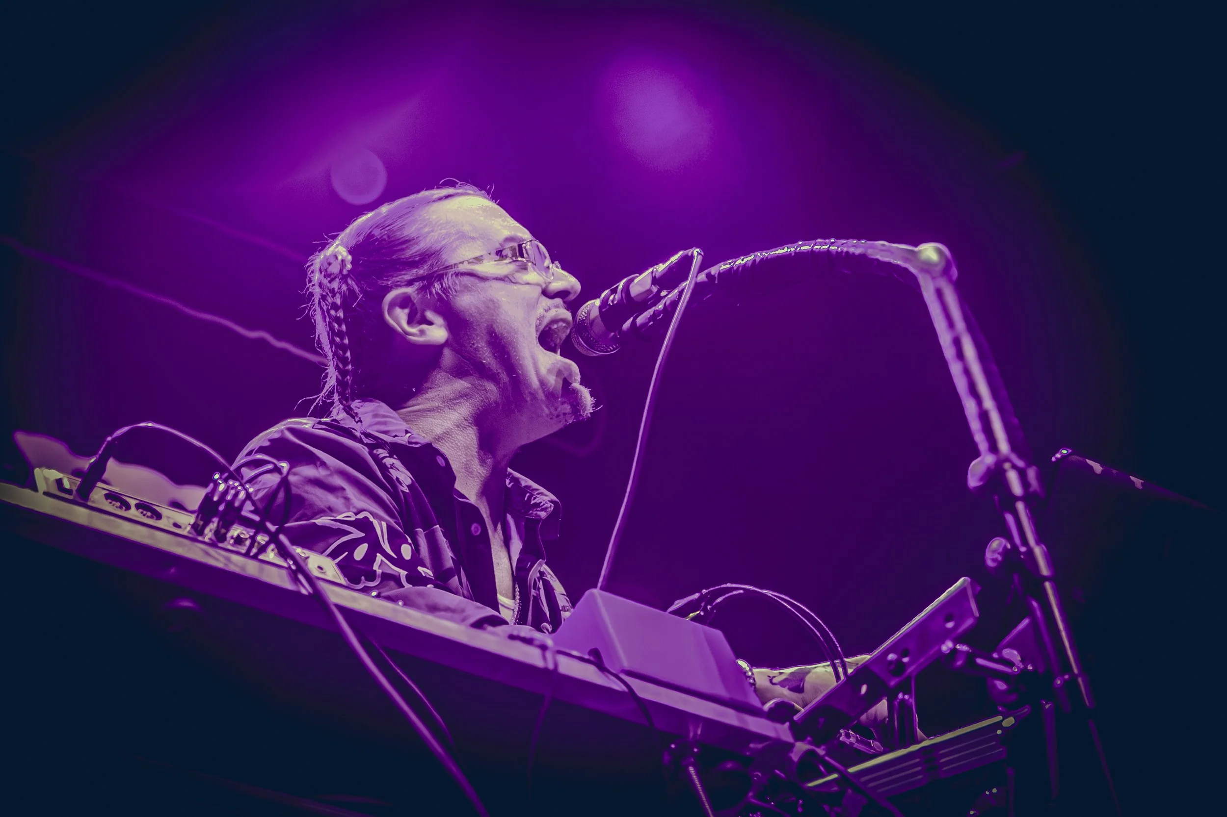 A musician with glasses and braided hair performing on stage with purple lighting, playing a keyboard and singing into a microphone.