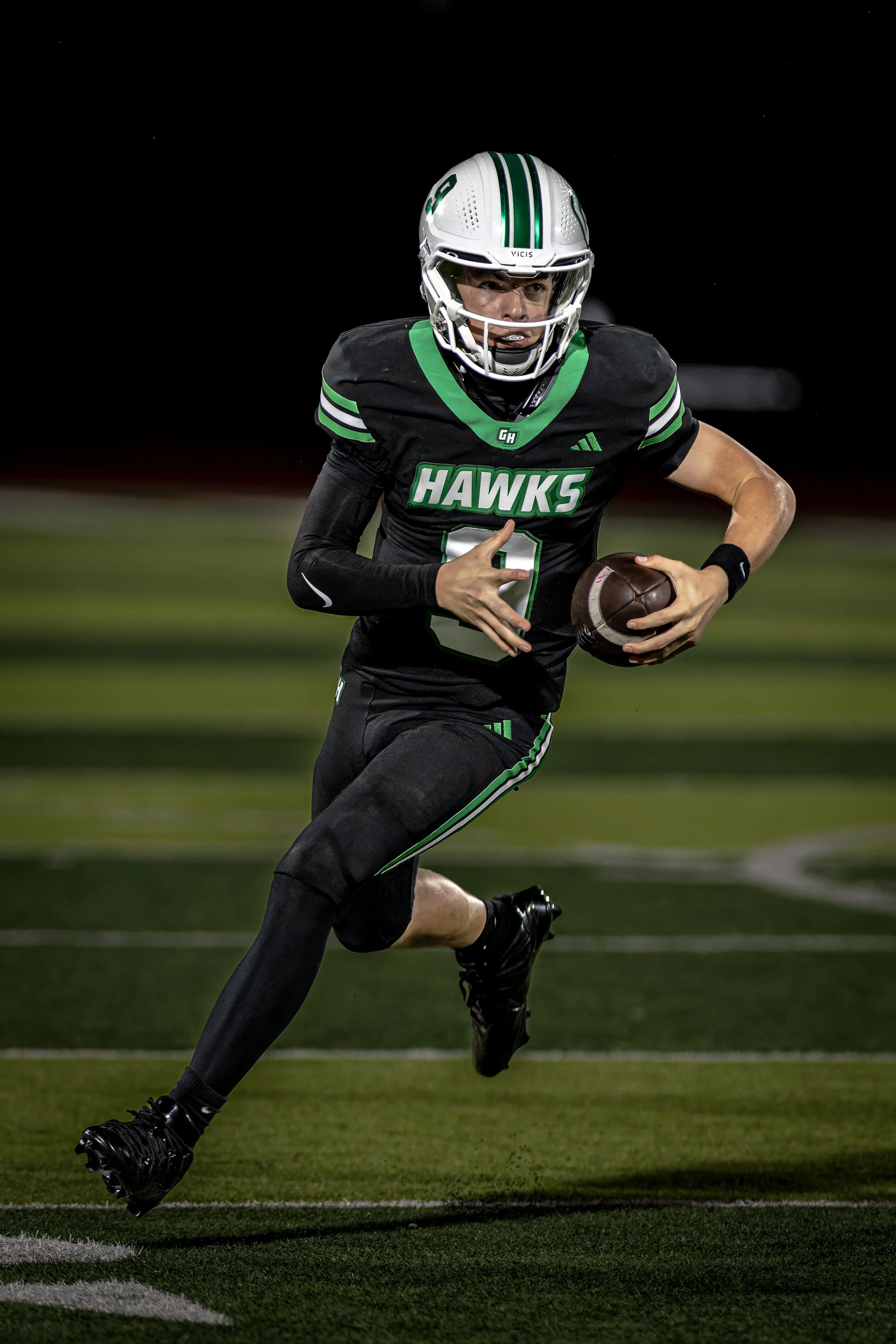 A football player in a black and green uniform running with the ball on a football field at night.