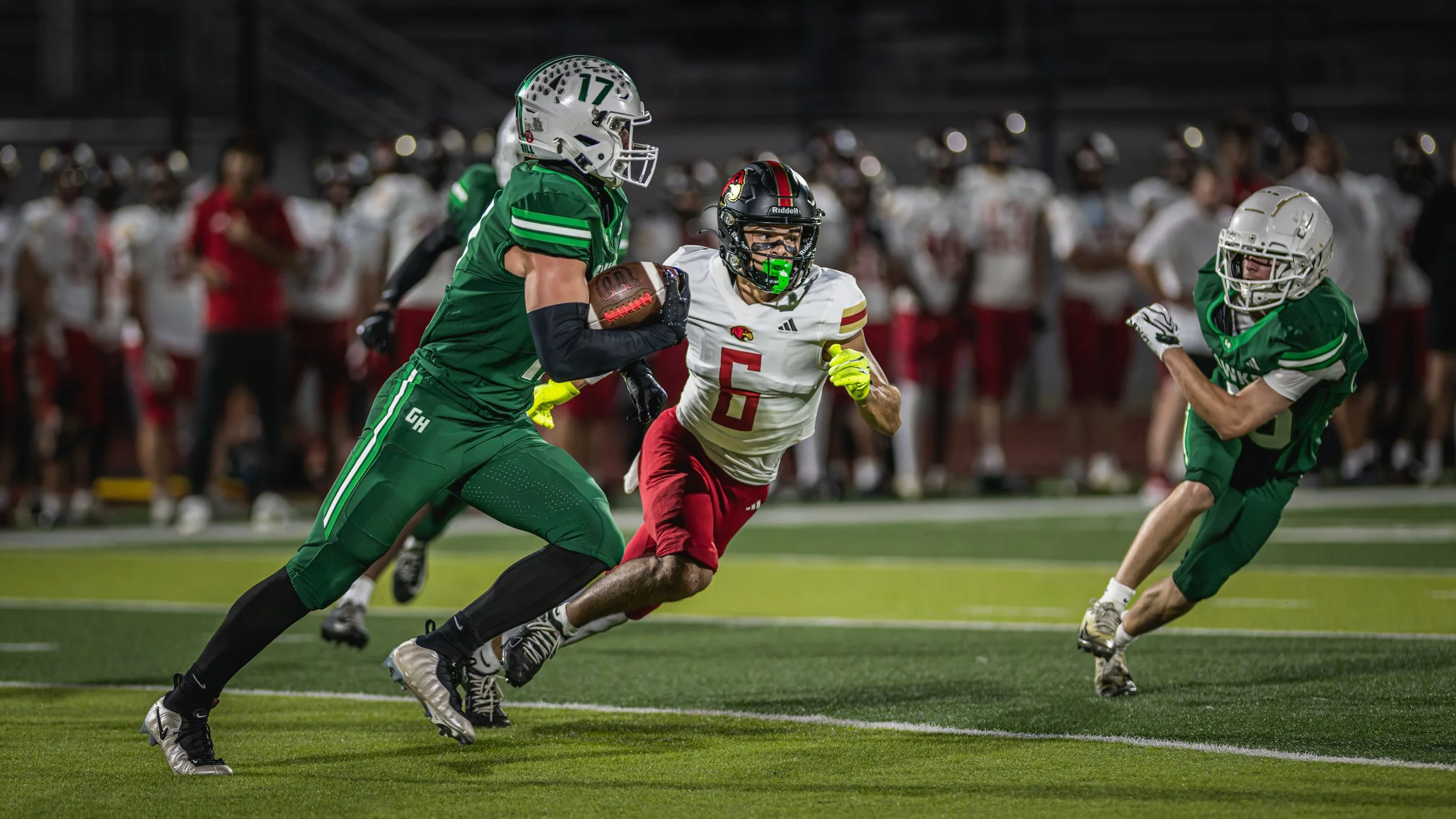 An American football game between two teams, one wearing green uniforms and the other in white and red. A player in green is running with the ball while a player in white and red attempts to tackle him. The scene takes place on a football field at ni