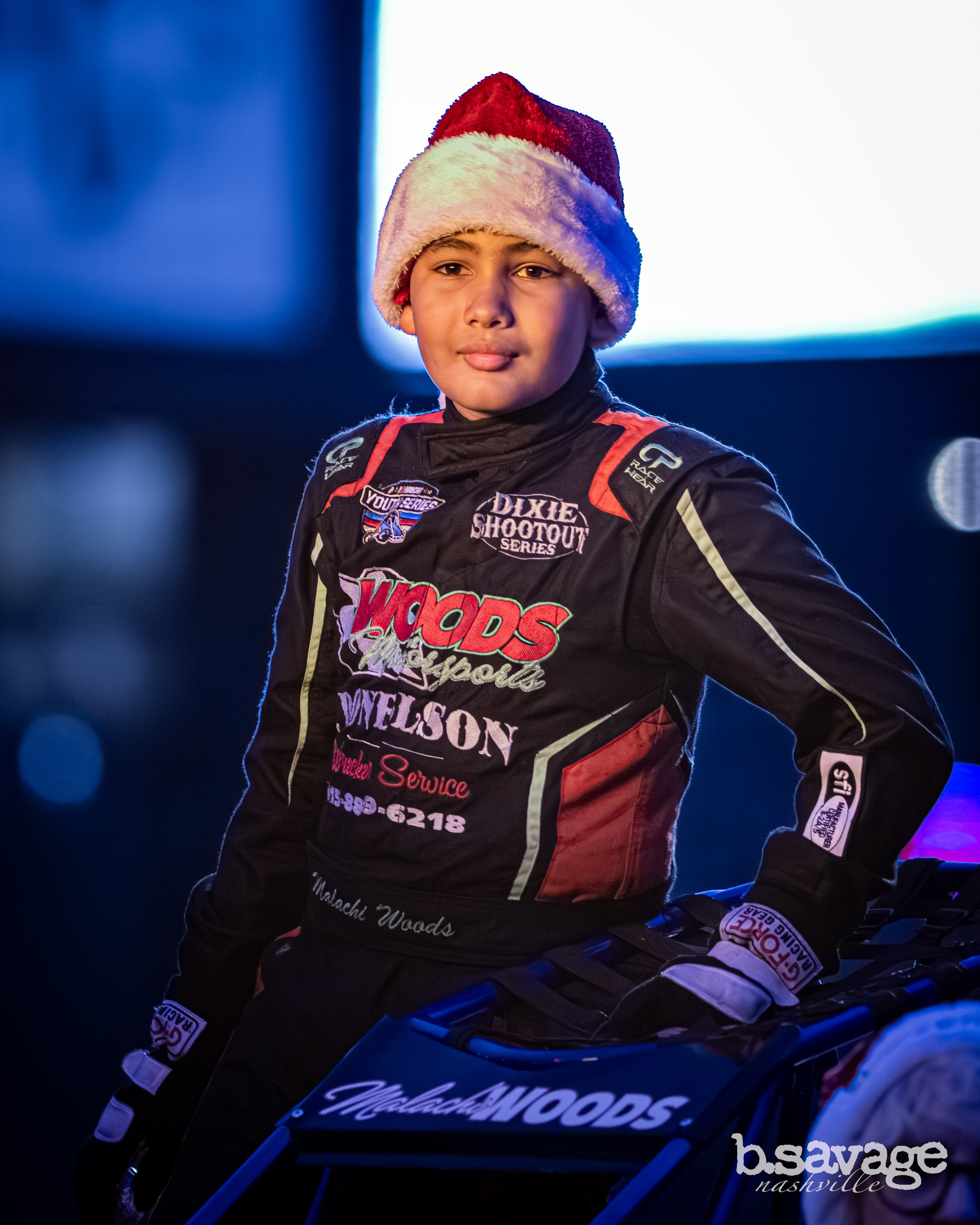 A young boy wearing a Santa hat and a racing suit with logos and sponsorship patches, sitting on a motorcycle at a race event.
