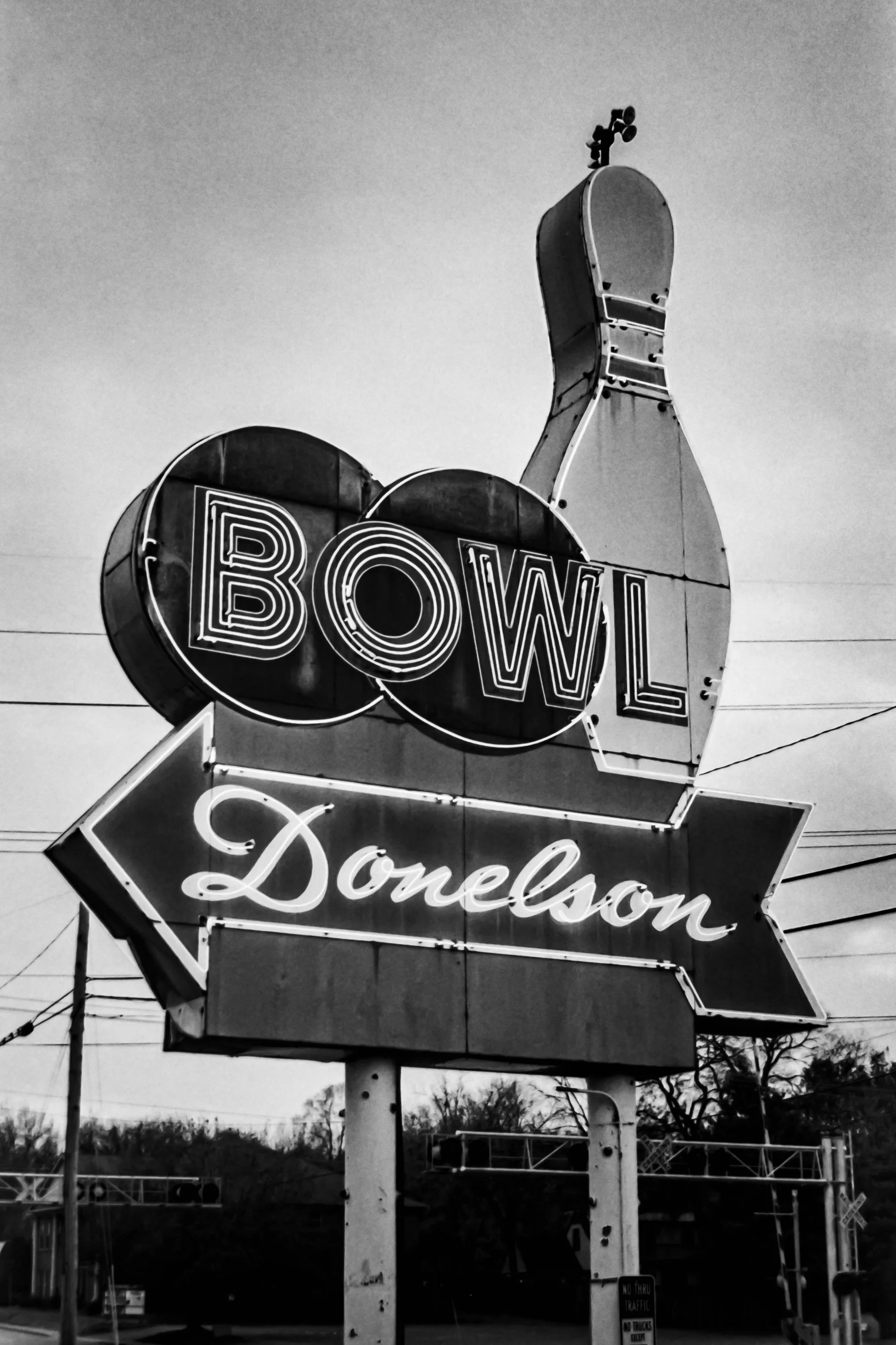 A vintage neon sign for Bowlero with the word 'Bowling' underneath, mounted on a tall pole, against a clear sky in black and white.