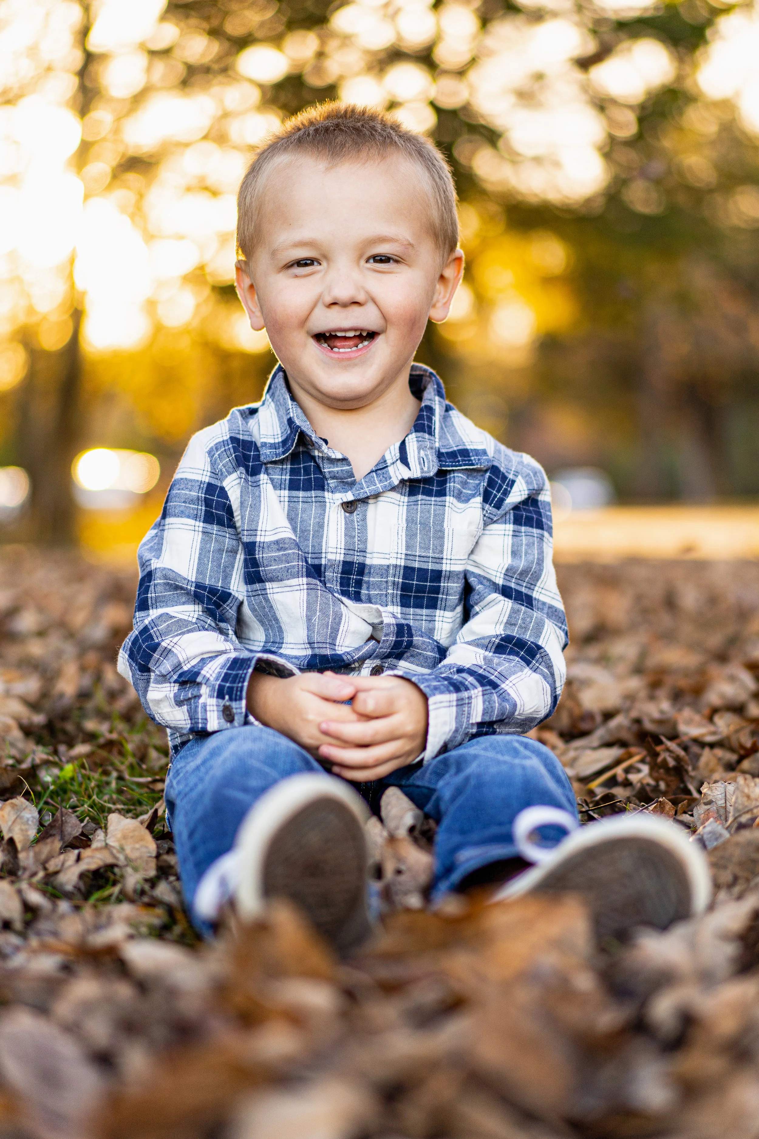 A young boy sitting on the ground outdoors during autumn, smiling at the camera, with fallen leaves, trees, and sunlight in the background.
