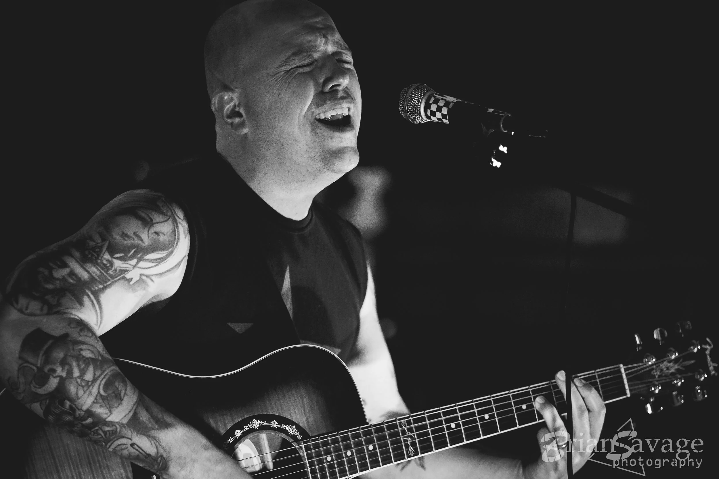 A man with tattoos singing passionately into a microphone while playing an acoustic guitar.