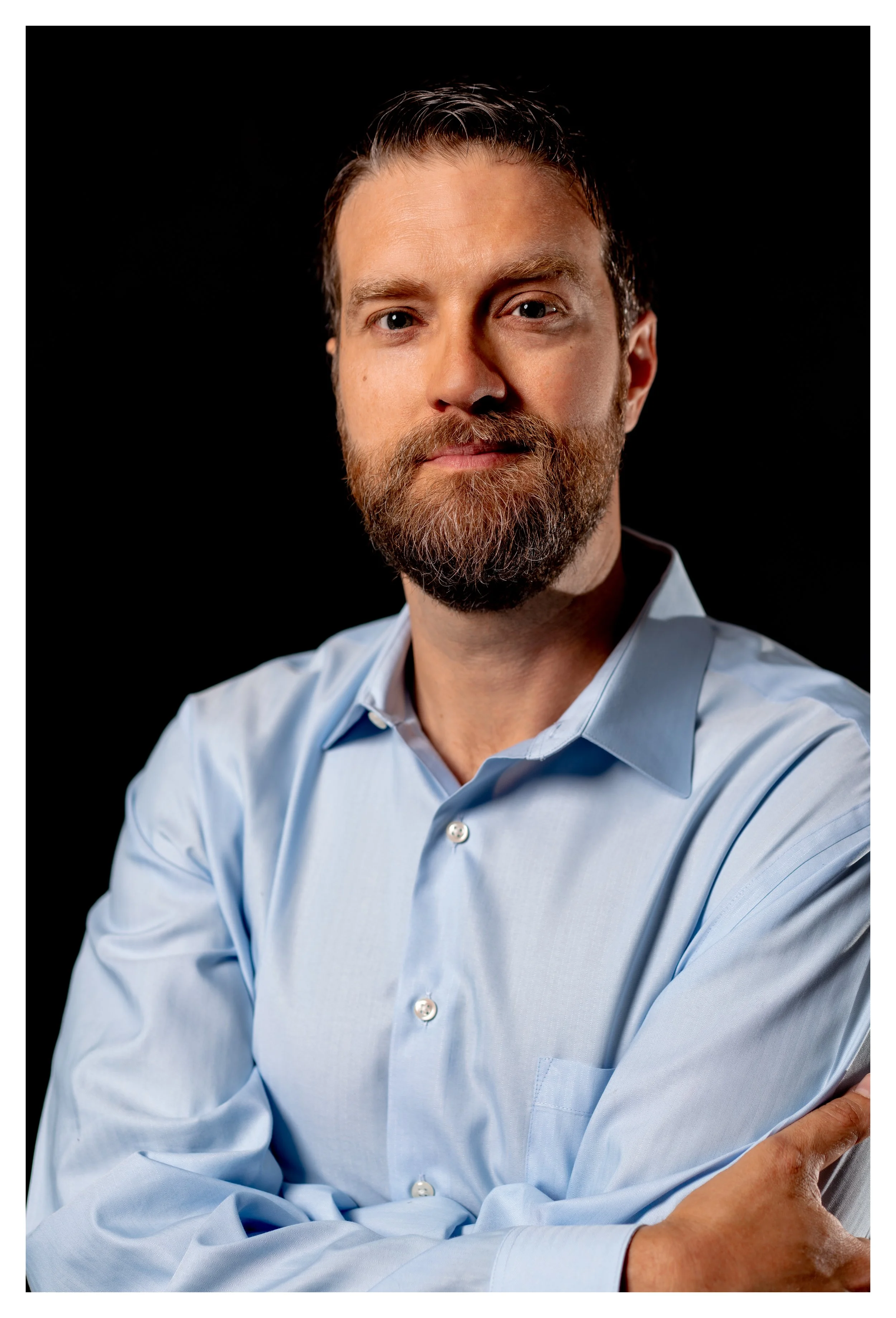 A man with light skin, brown hair, and a beard, wearing a light blue dress shirt, looking at the camera with a neutral expression, against a black background.
