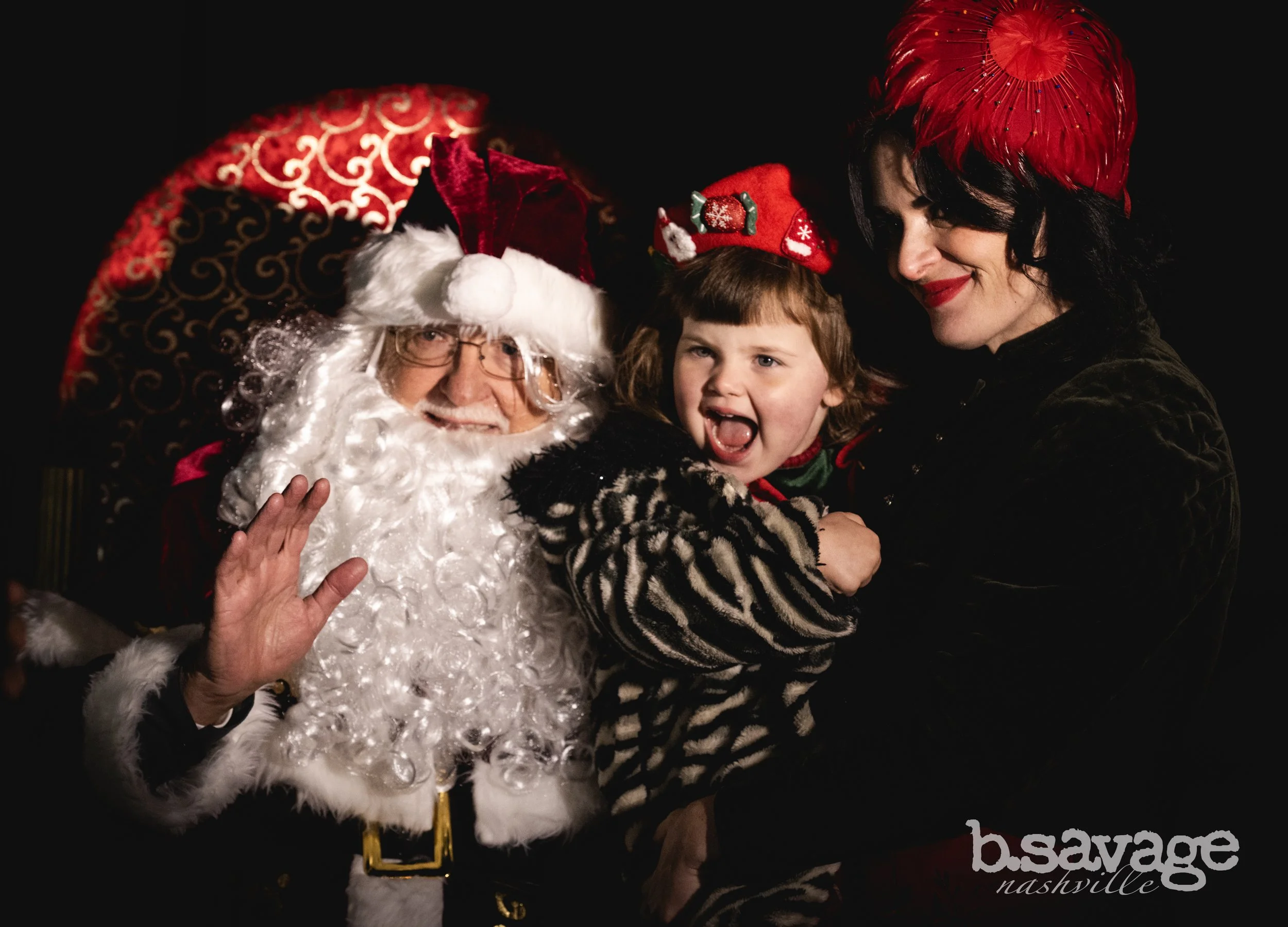 A woman, a man dressed as Santa Claus, and a young girl smiling and hugging each other against a dark background, with Christmas hats and festive clothing.