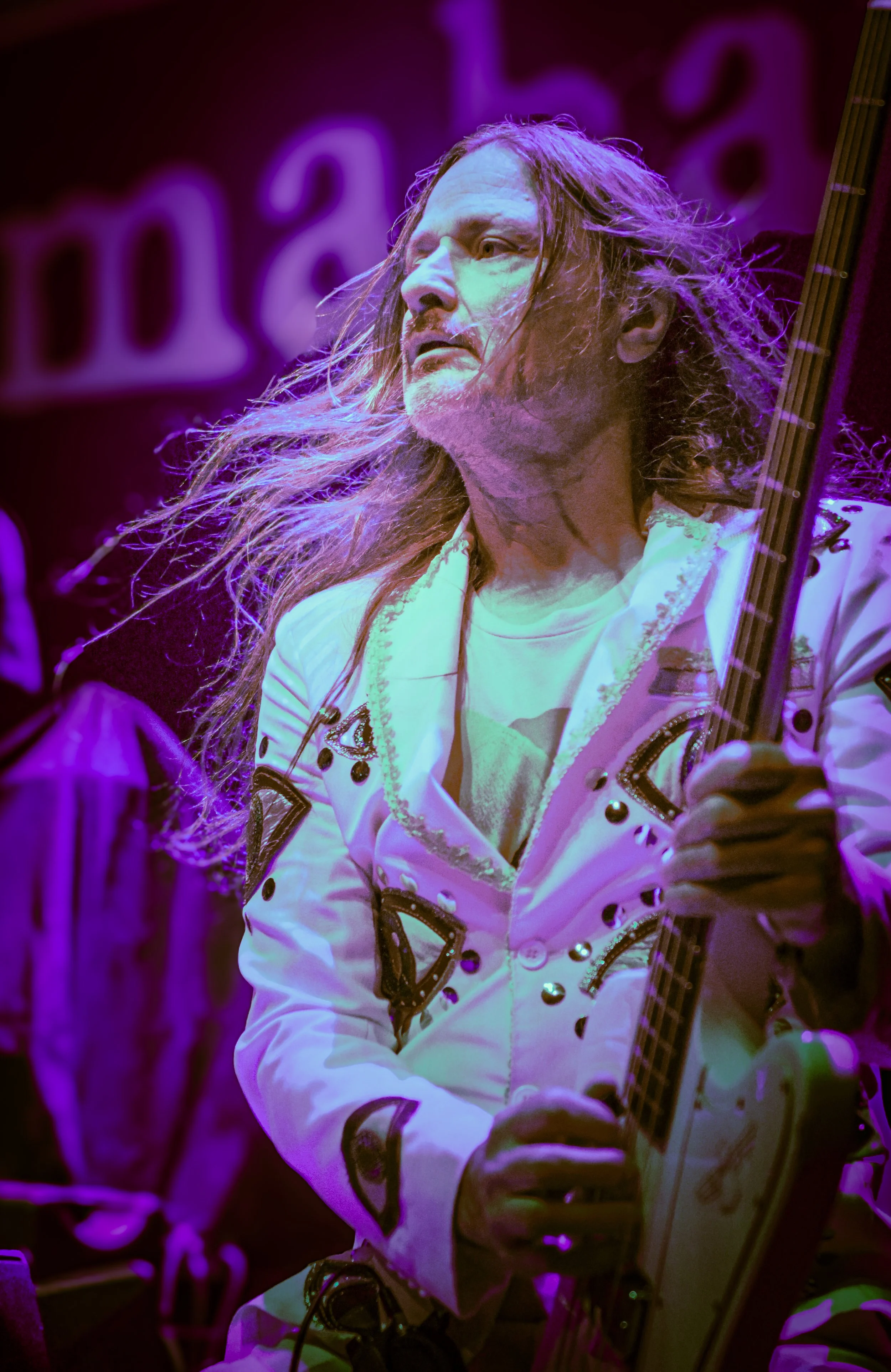 A man with long, wavy hair and a mustache playing an electric guitar on stage illuminated by purple and pink lights.