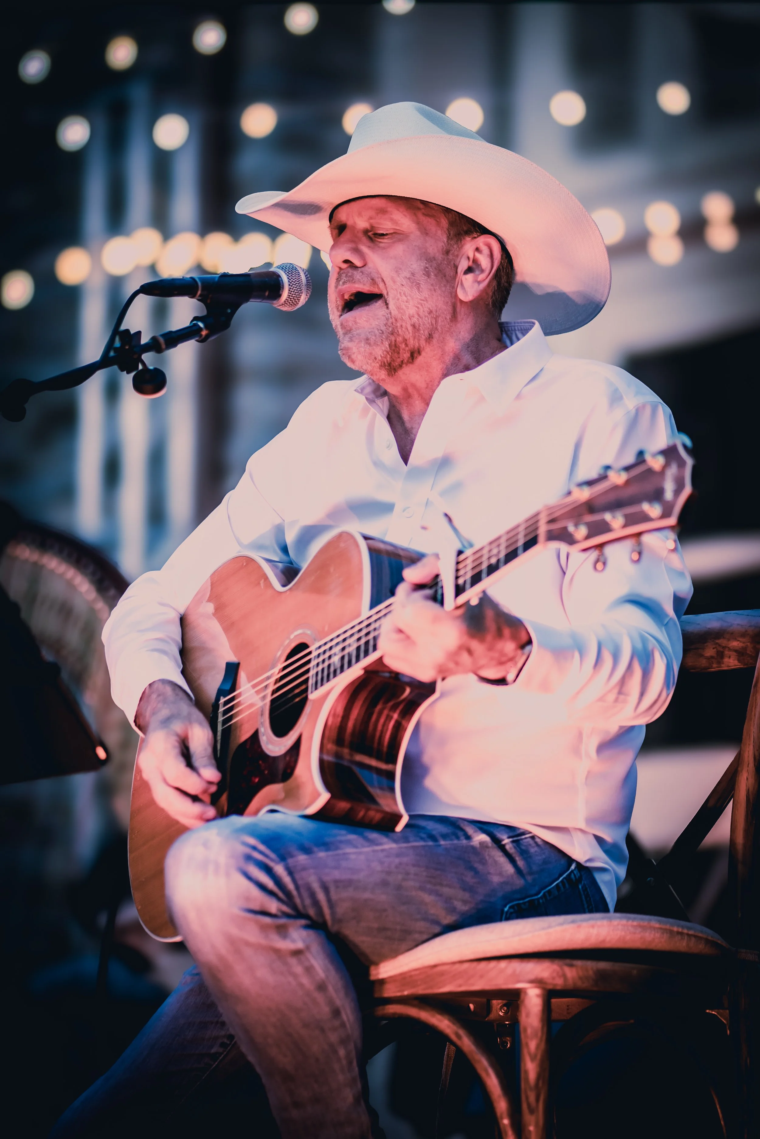 A man wearing a cowboy hat and white shirt, playing an acoustic guitar and singing into a microphone on stage at night, with blurred string lights in the background.