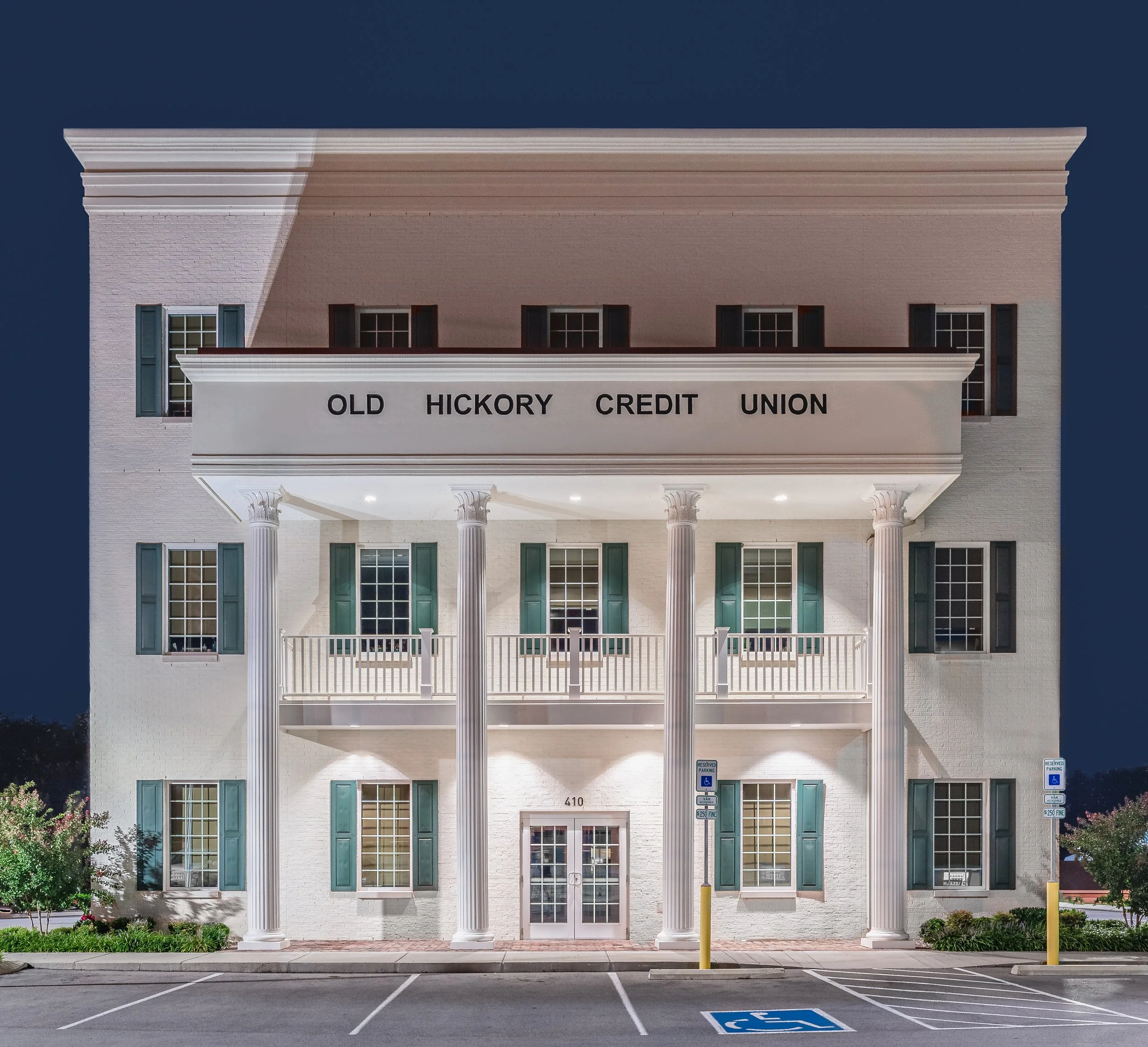 The facade of a white building with green shutters, columns, and a balcony labeled 'Old Hickory Credit Union.' There are parking spaces in front, including a reserved handicapped space.