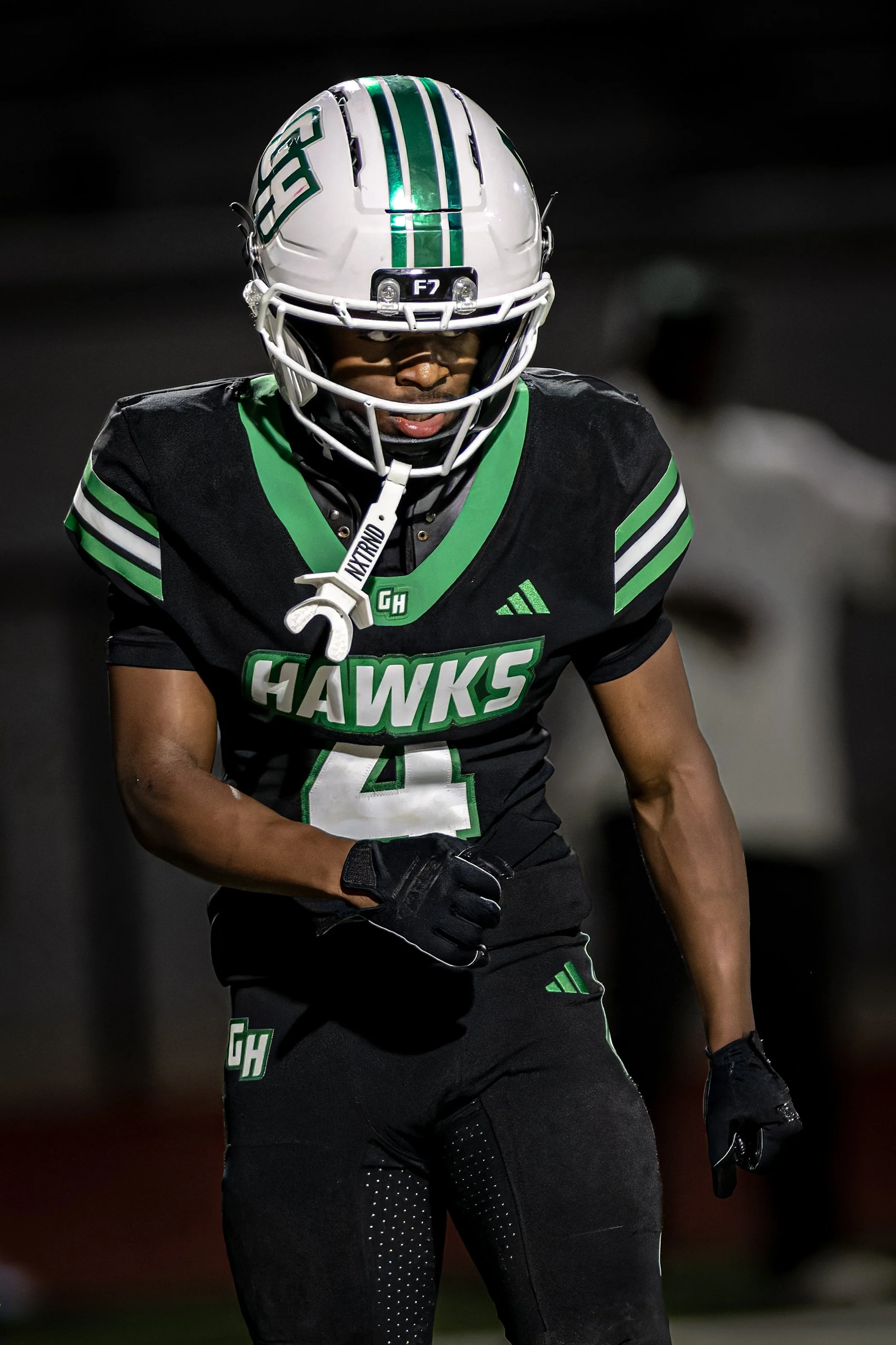 A football player in black and green uniform with a white helmet and face shield, captured during a game at night.