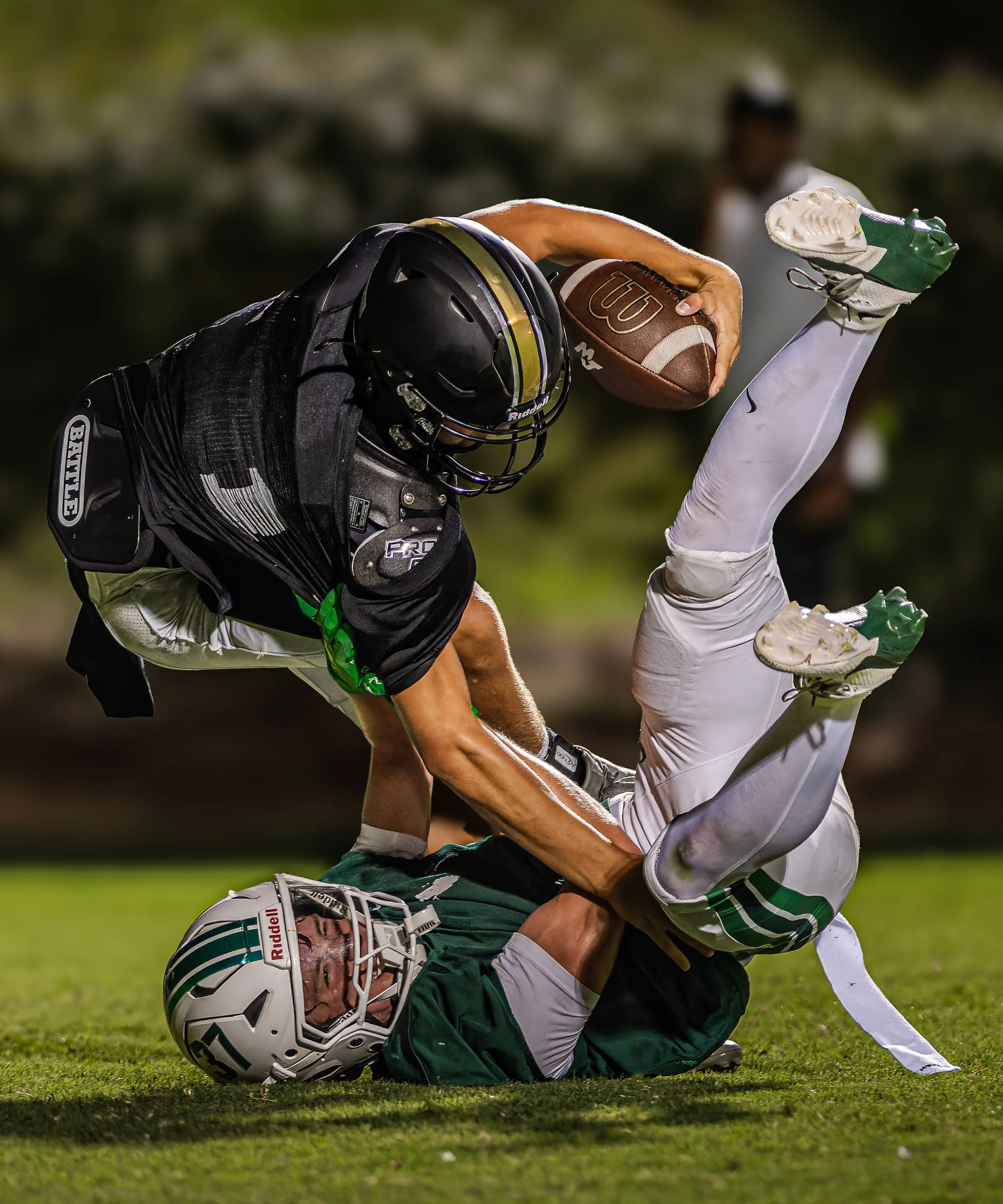An intense moment during a football game where a player in black and green gear is tackling a player in white and green gear on the field at night.