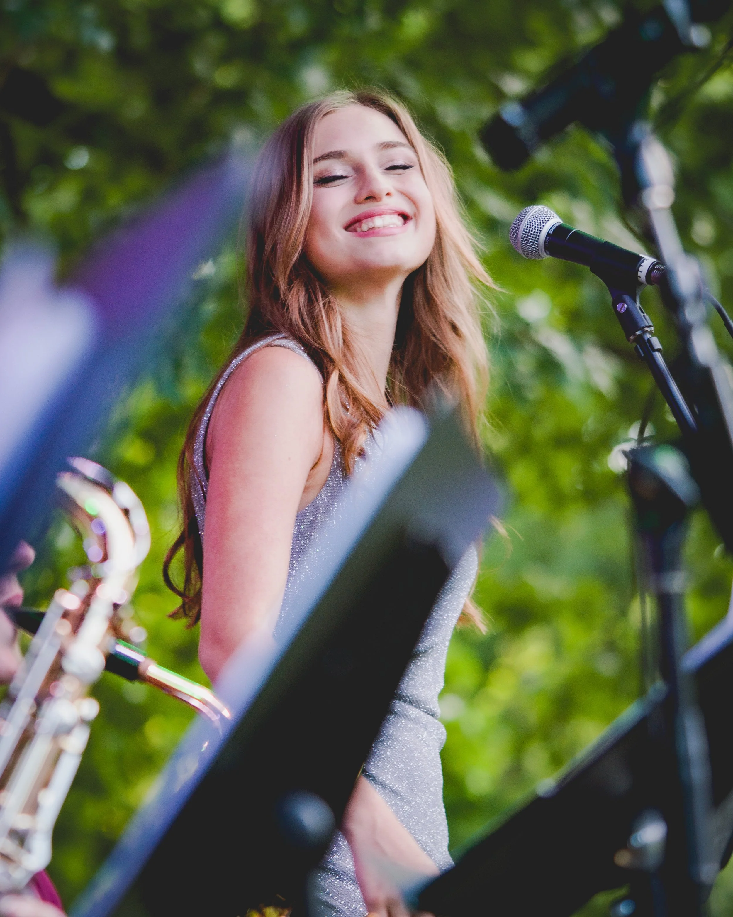 A young woman with long, wavy light brown hair smiling with eyes closed, singing into a microphone outdoors with green trees in the background.
