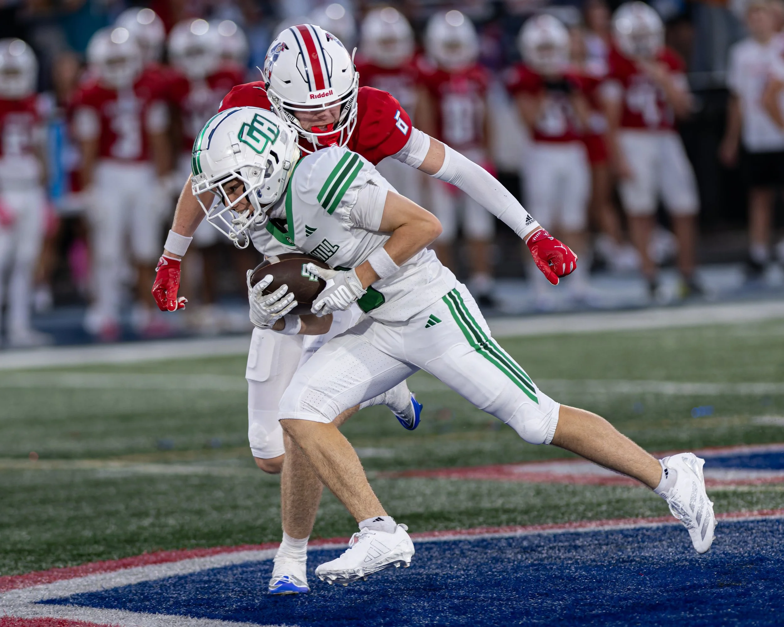 Two football players in action on the field: one in a white uniform with green accents, holding a football, and the other in a red uniform tackling him. The player in white is bent over with his head down, and the player in red is reaching over him. 