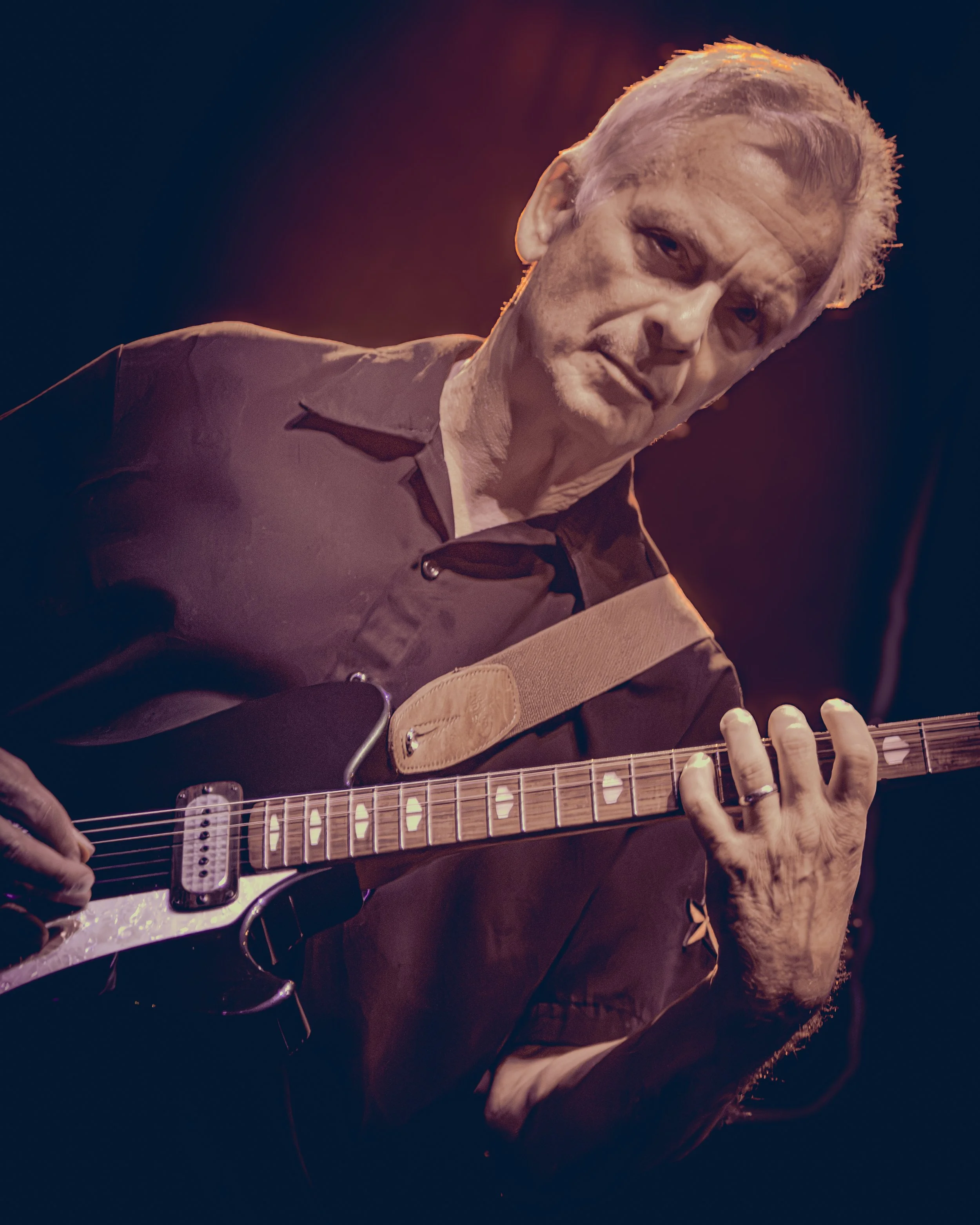 A person playing an electric guitar against a dark background with a reddish glow, wearing a dark jacket and rings on their fingers.